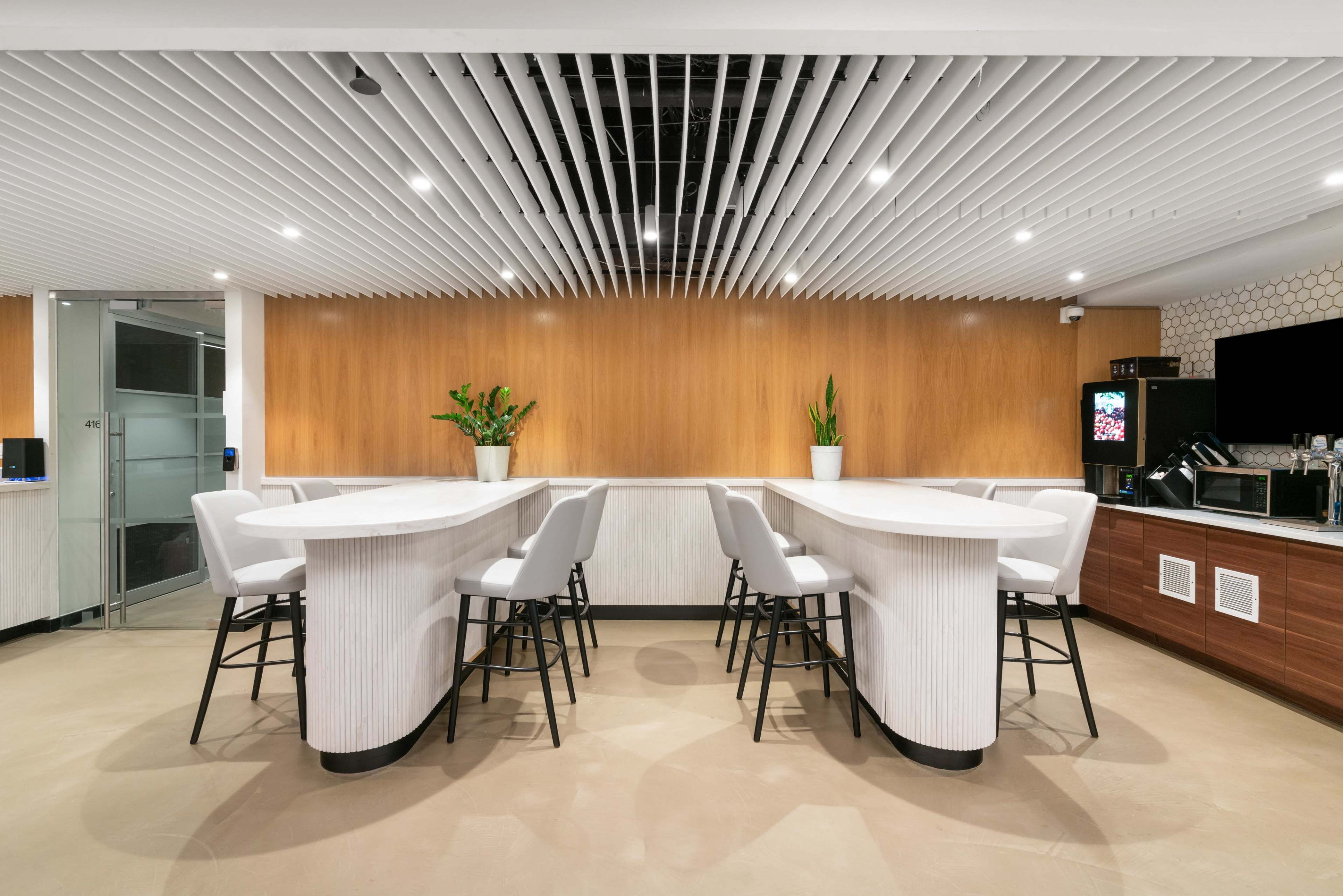 The image shows a modern, minimalist lounge area featuring a curved white bar table surrounded by high chairs, with wooden paneling and plants in the background.