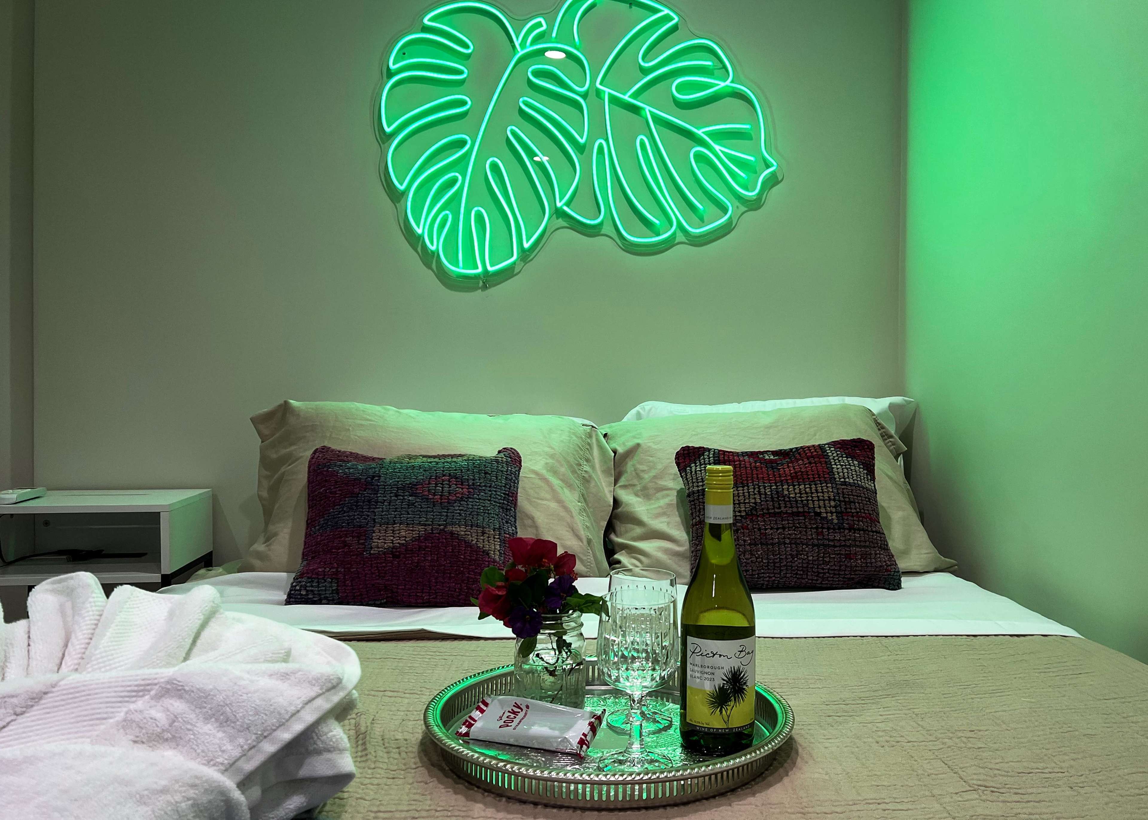 A neatly arranged bed is accented by a neon green leaf sign, with a tray featuring a bottle of white wine, glasses, and a small floral decoration.
