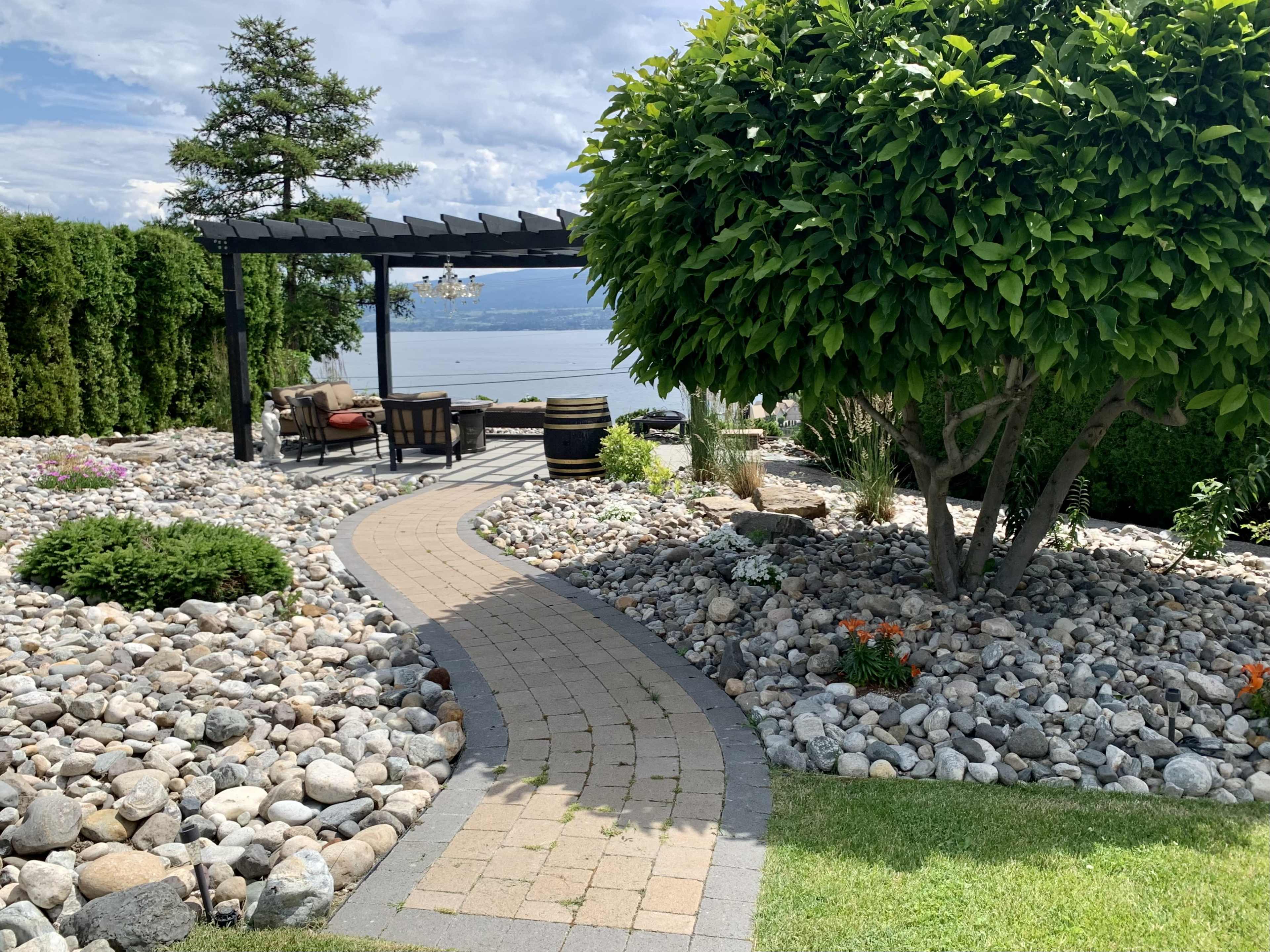 A winding stone pathway leads through a landscaped garden with rocks, shrubs, and a seating area overlooking a body of water.