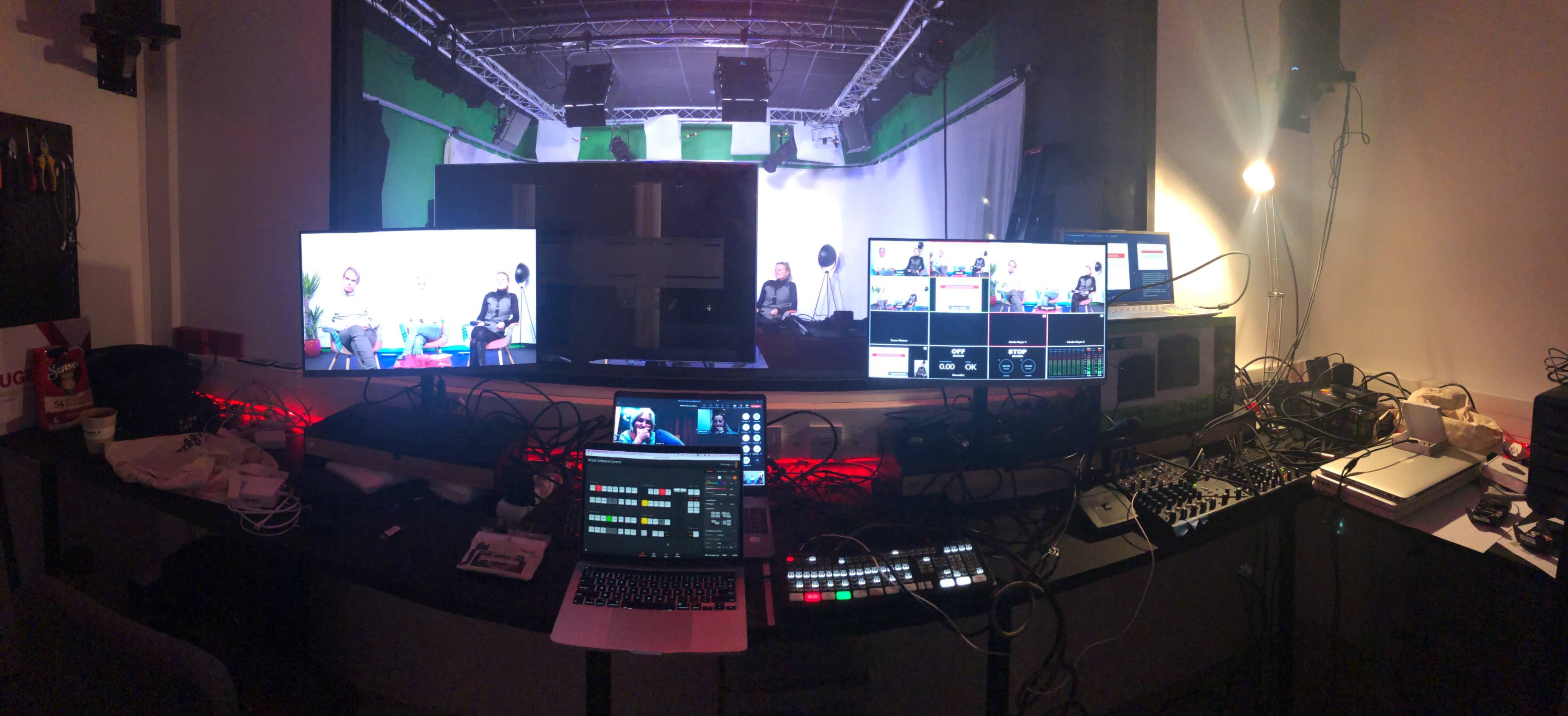 A control room with multiple screens displaying a live video feed, along with various electronic equipment and cables on a desk.