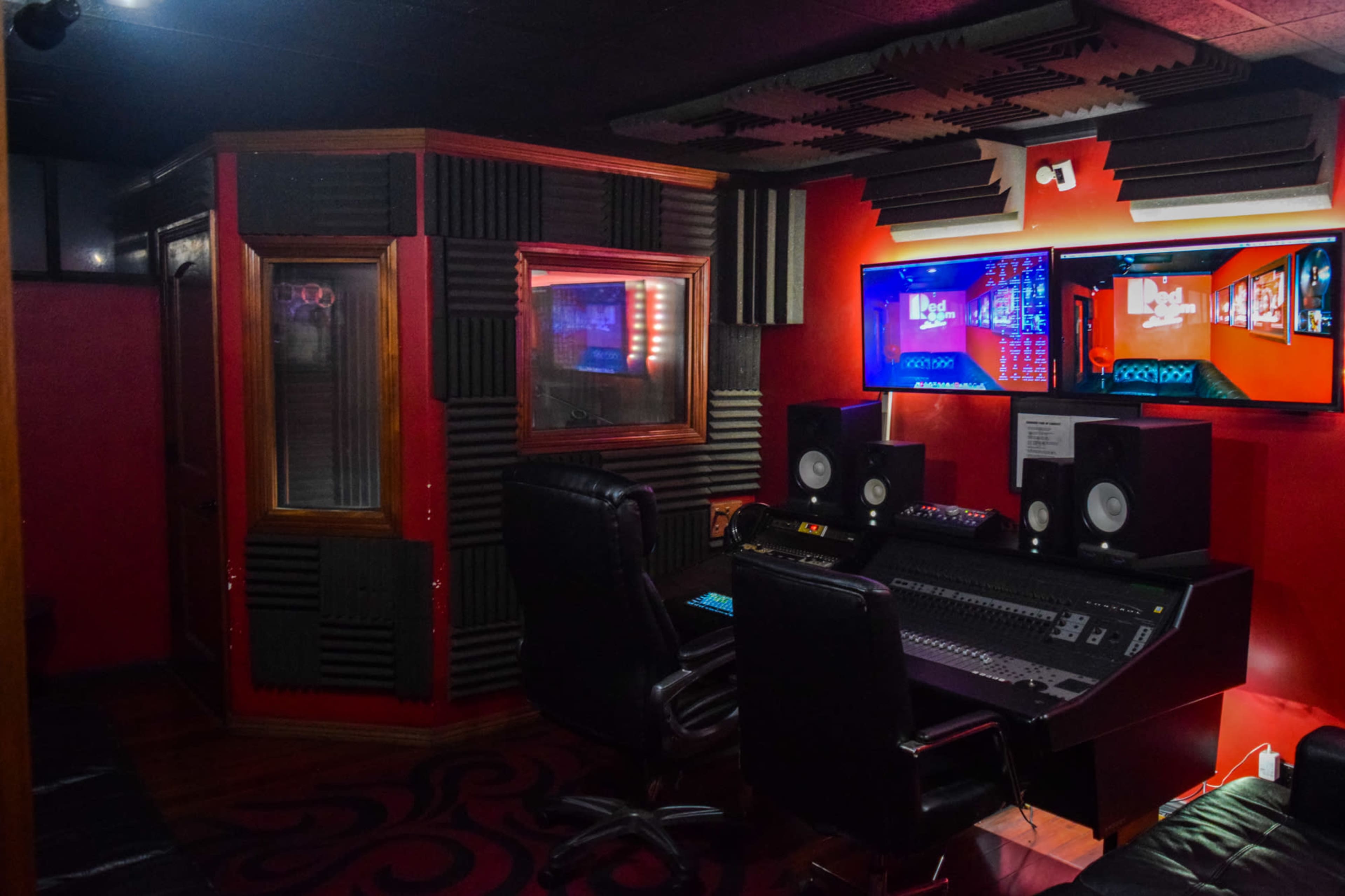 The image shows a music studio featuring a soundproof booth, a mixing console, and two large monitors on a red wall.