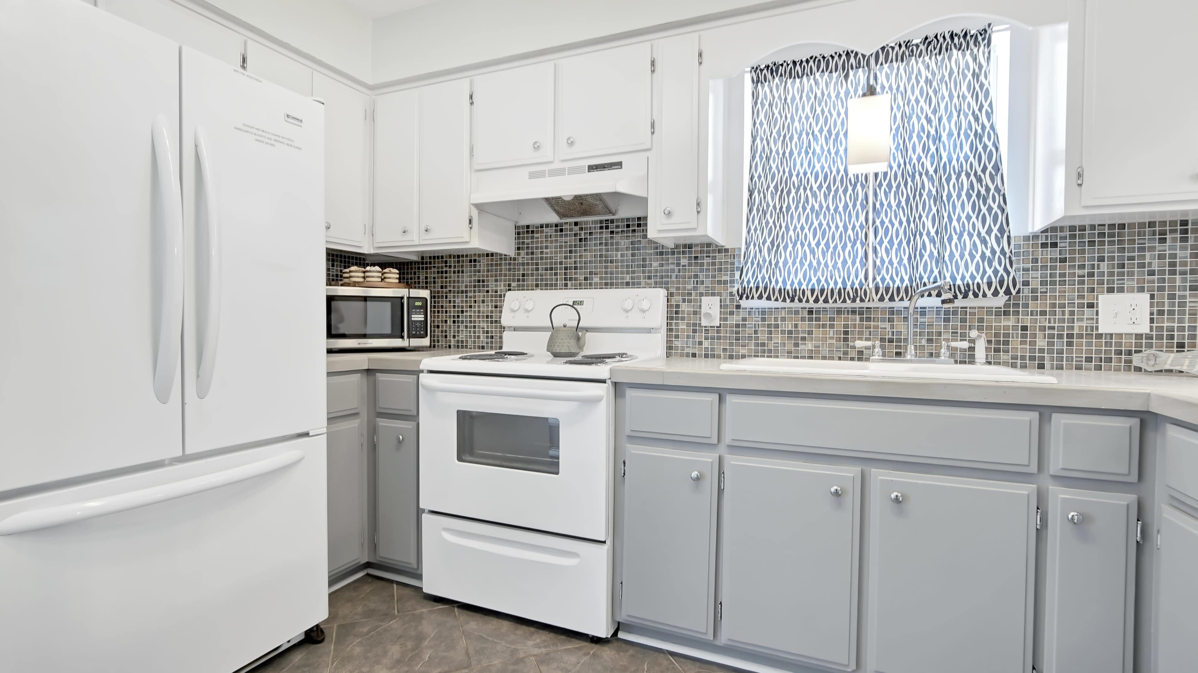 A modern kitchen features white cabinets, a gray countertop, and a stove with a microwave, along with a window dressed in a patterned curtain.