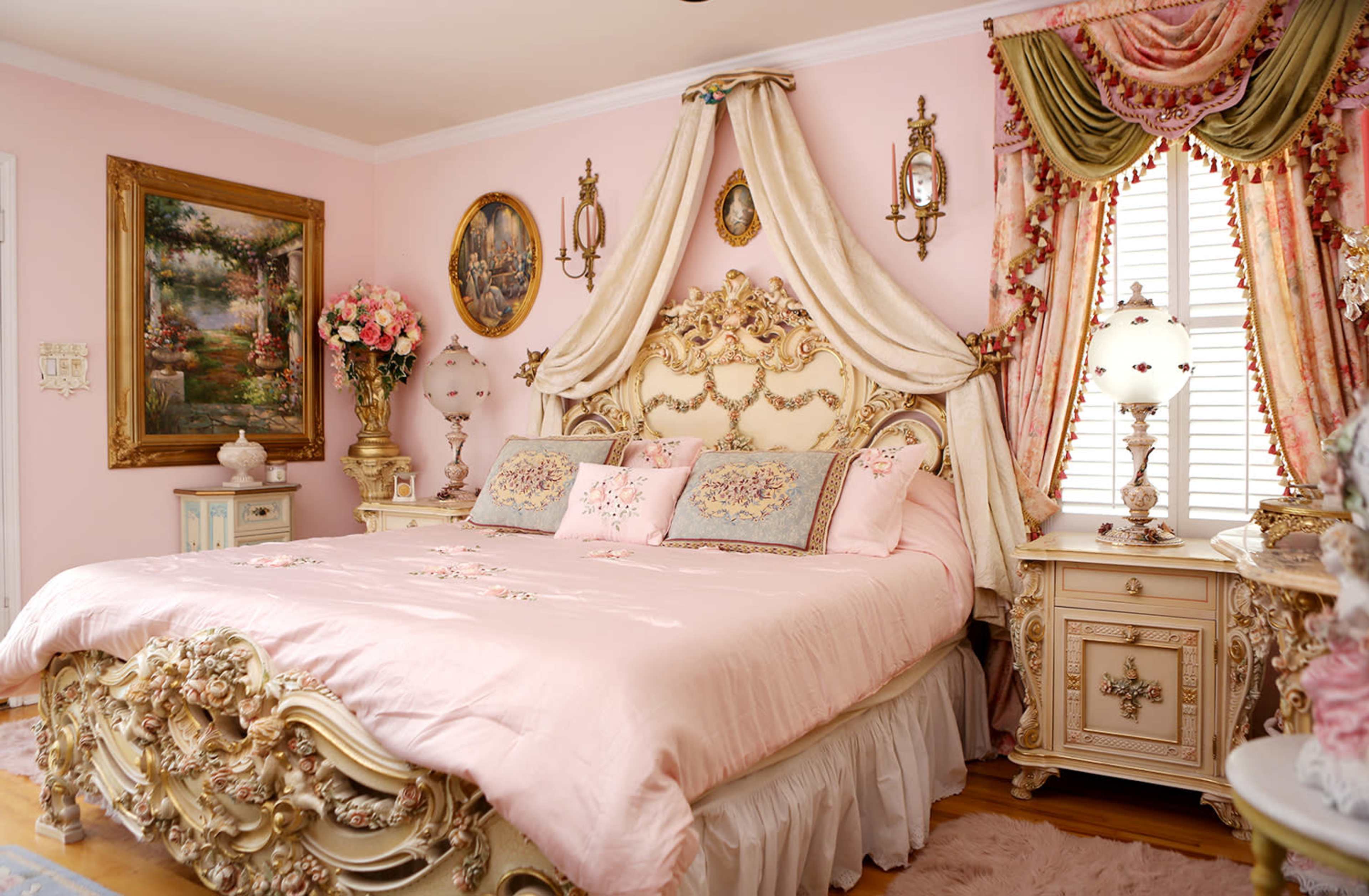 A lavishly decorated bedroom with a large, ornate bed covered in pink bedding, framed art on the walls, and elegant curtains draped over the windows.
