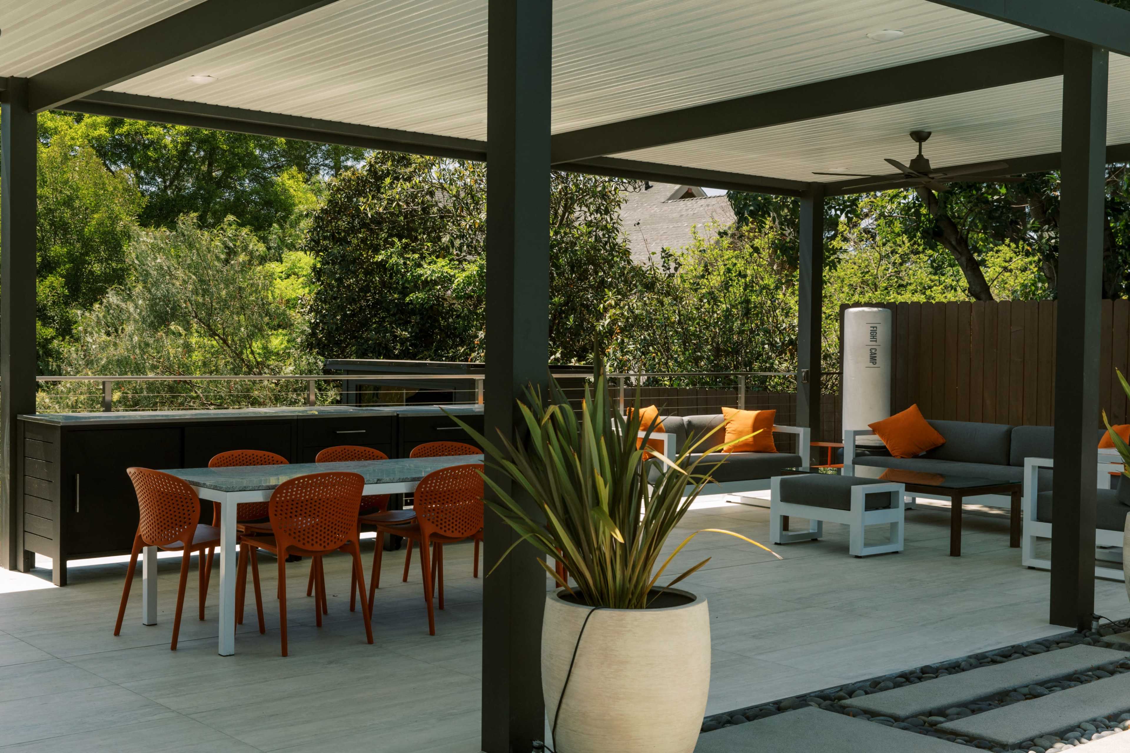 A modern outdoor patio features a dining table with orange chairs, seating areas, and greenery surrounding the space.