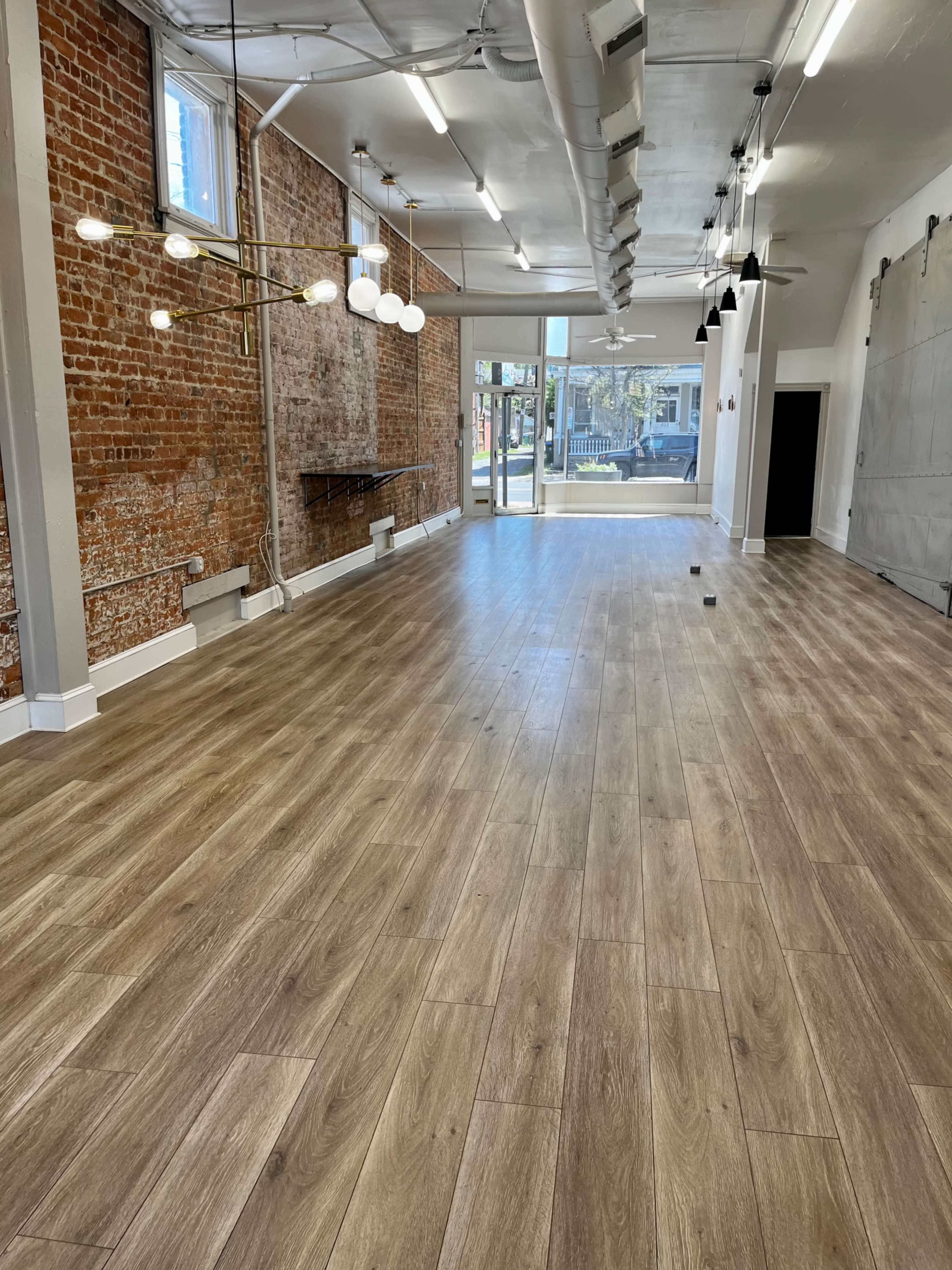 A spacious room with wooden flooring, exposed brick walls, and large windows allowing natural light, featuring modern light fixtures and ceiling fans.