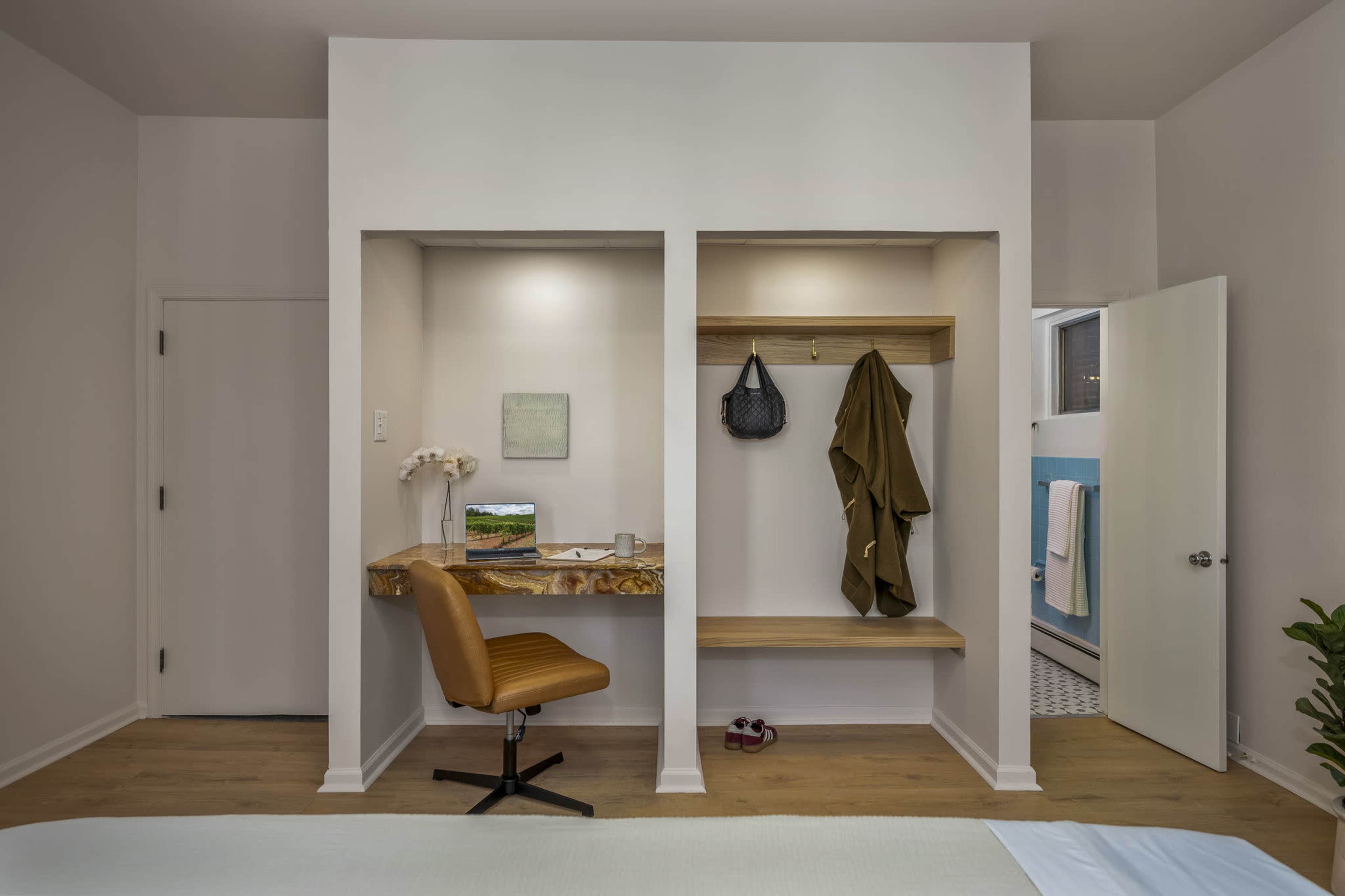 The image shows a minimalist workspace with a wooden desk, a chair, and a coat rack in a well-lit room.