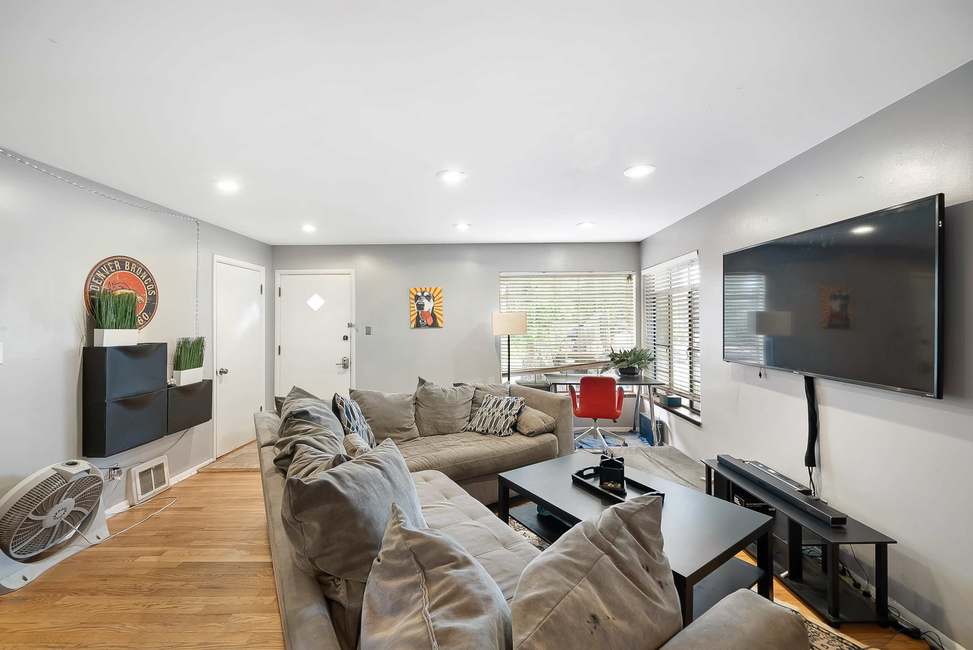 The image shows a living room with a large sectional sofa, a coffee table, and a wall-mounted television, featuring modern decor and ample natural light from windowed walls.
