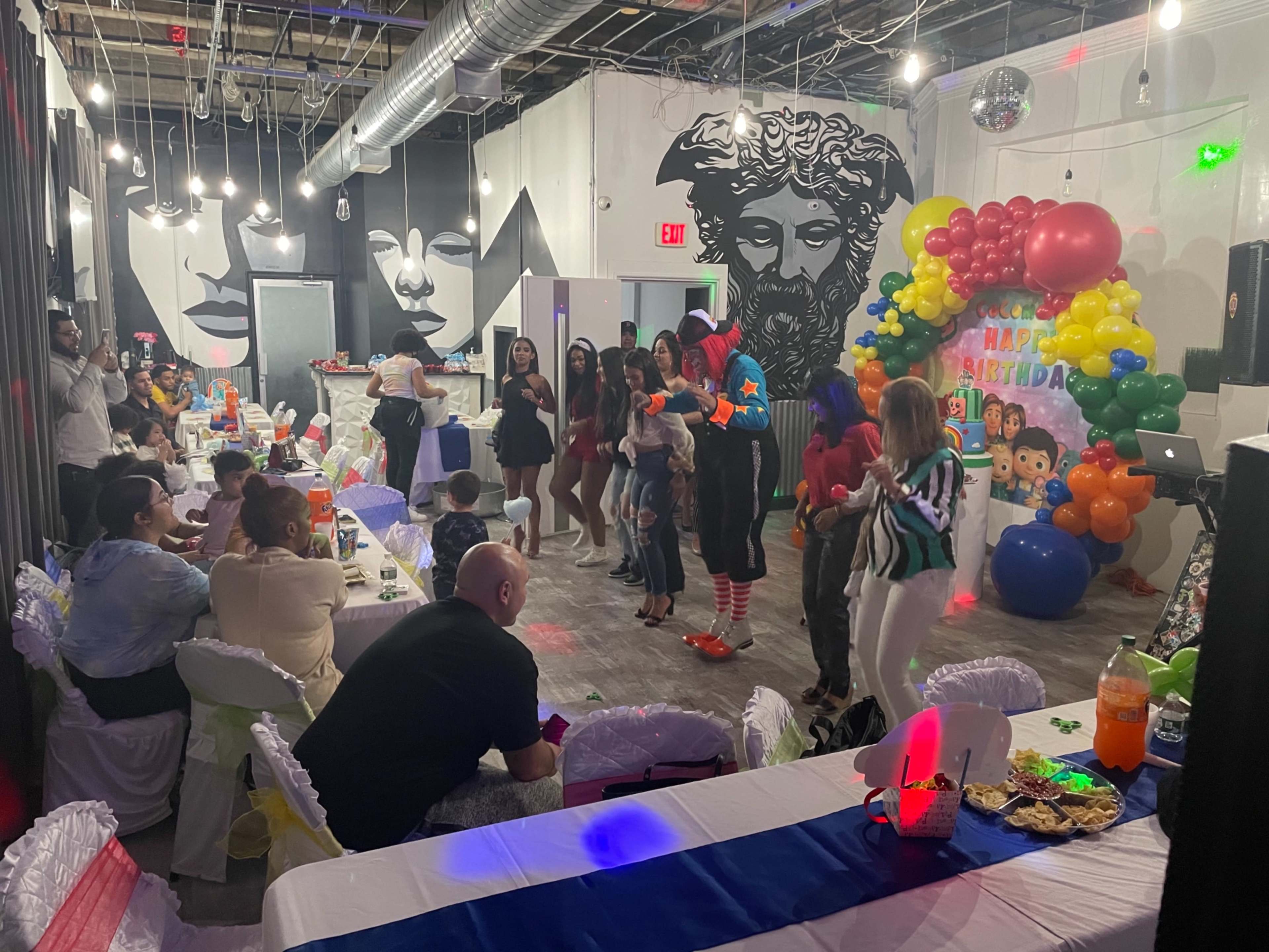 The scene depicts a colorful birthday party with guests seated at tables while others dance in front of a festive backdrop featuring balloons and party decorations.