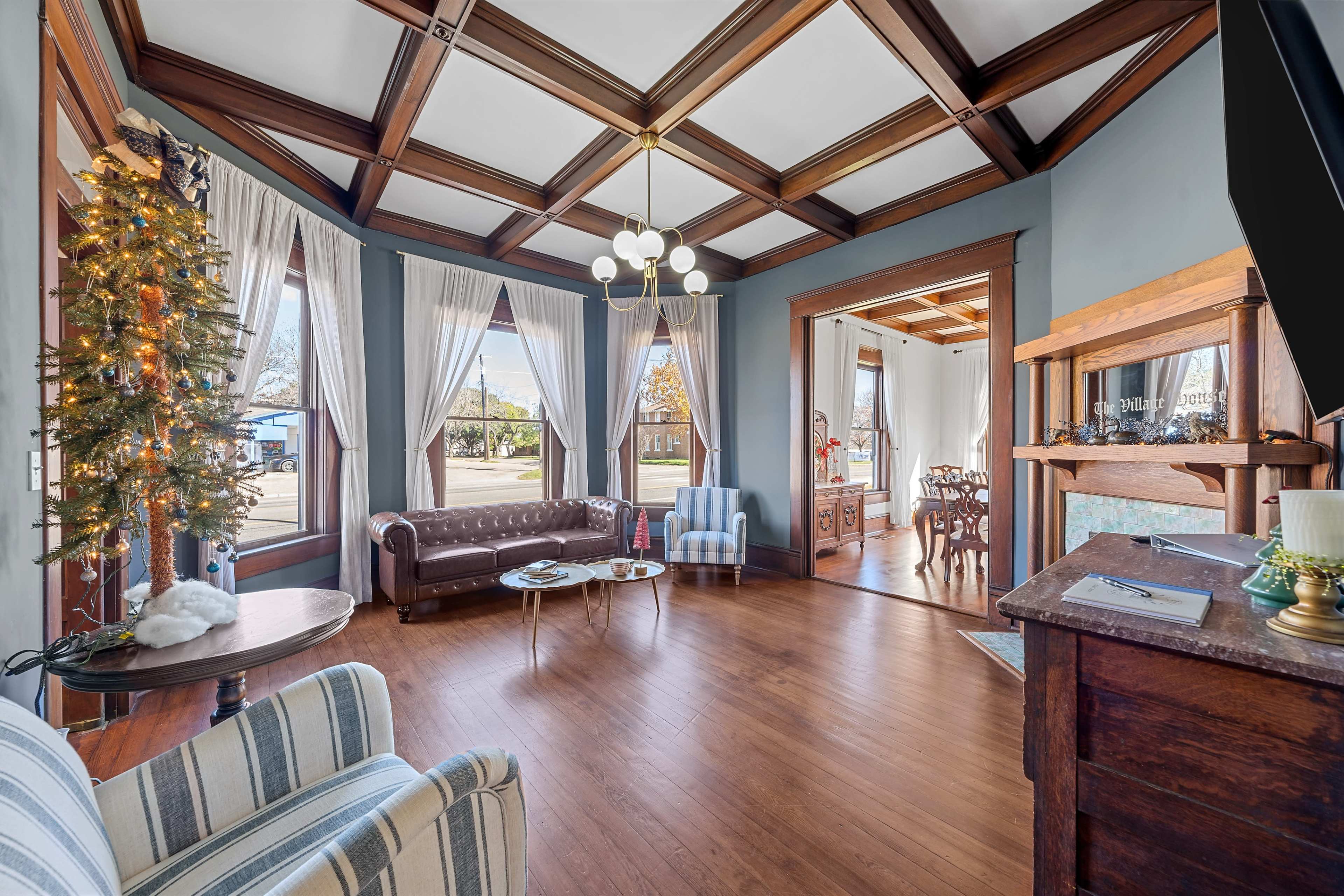 A spacious living room features a coffered ceiling, elegant windows with drapes, and a decorative Christmas tree near a leather sofa and wooden furniture.
