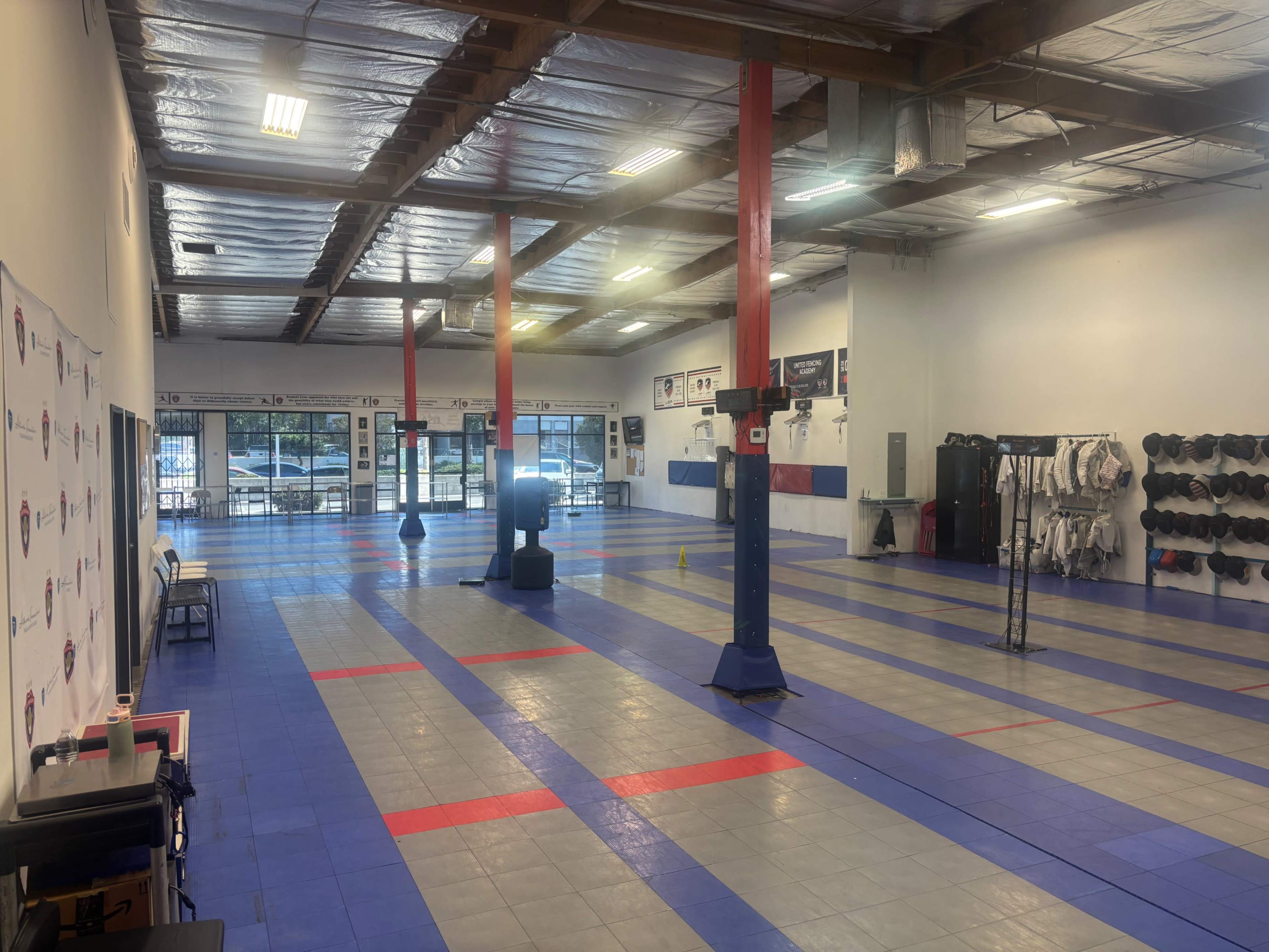 The image shows a spacious martial arts training area with mats, a training dummy, and fitness equipment along the walls, illuminated by overhead lights.