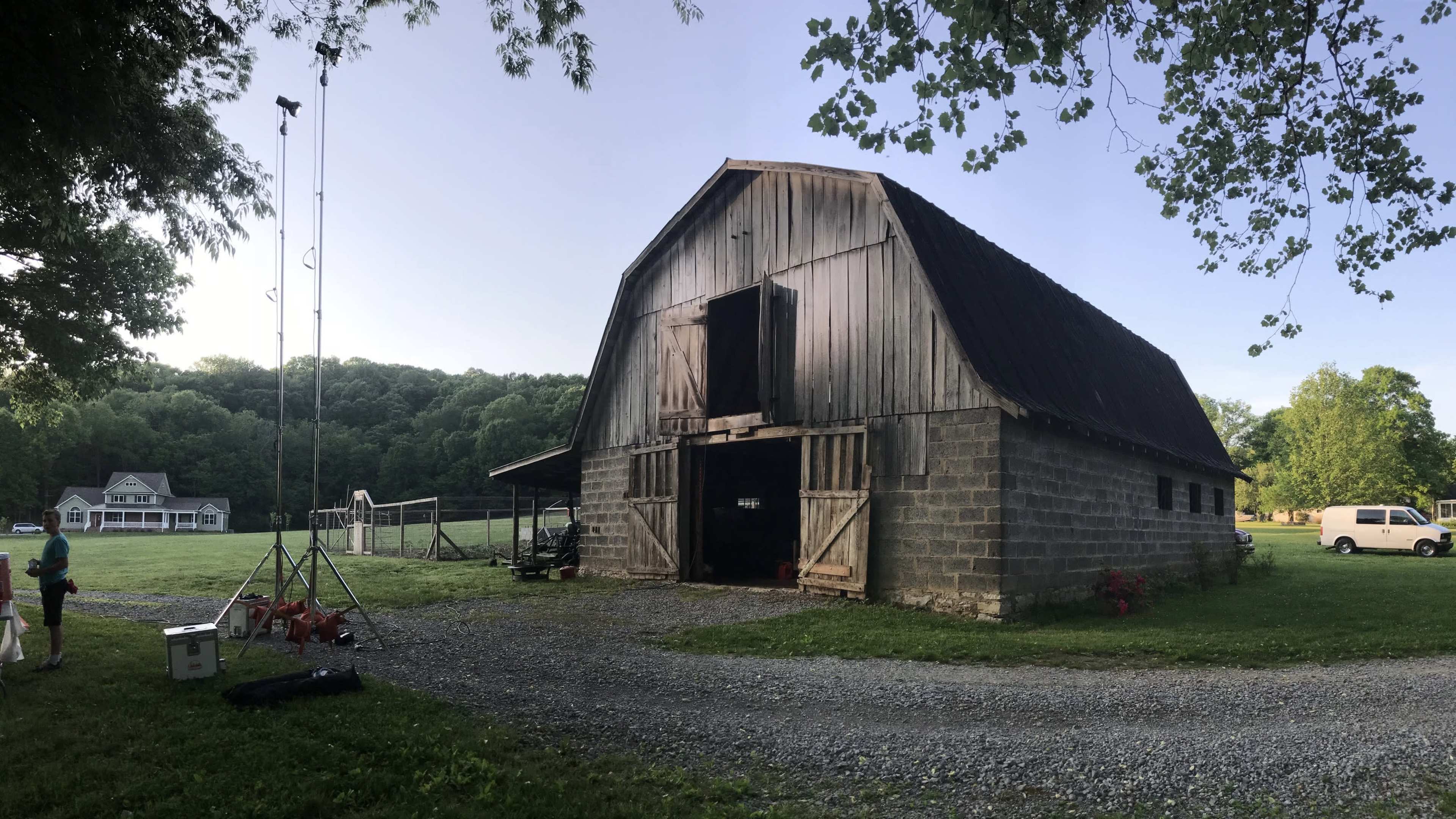 A large wooden barn with a metal roof stands beside a gravel path, surrounded by greenery and a field in the background.