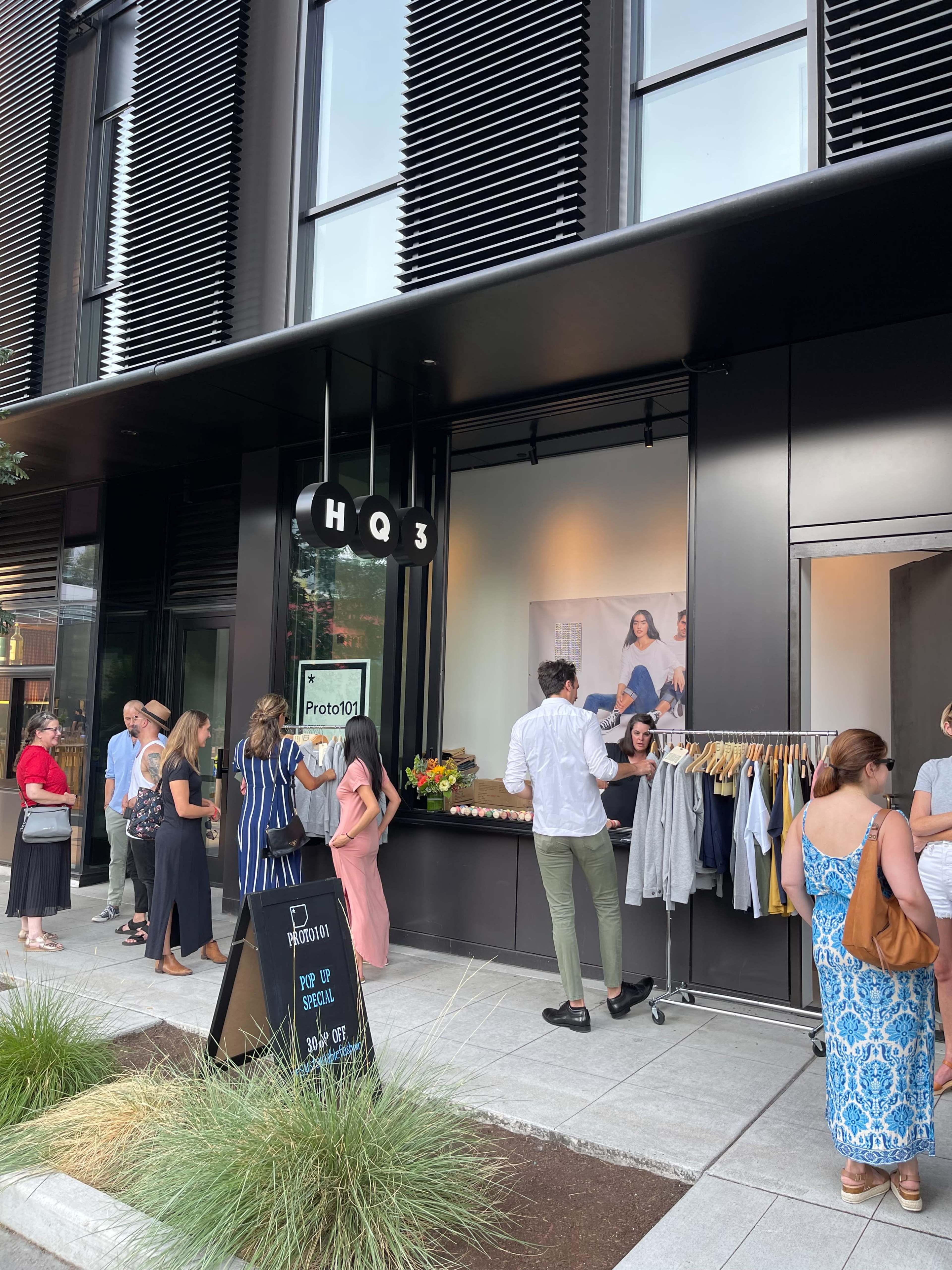 A crowd of people gathers outside a modern retail space featuring a display of clothing and a promotional sign.