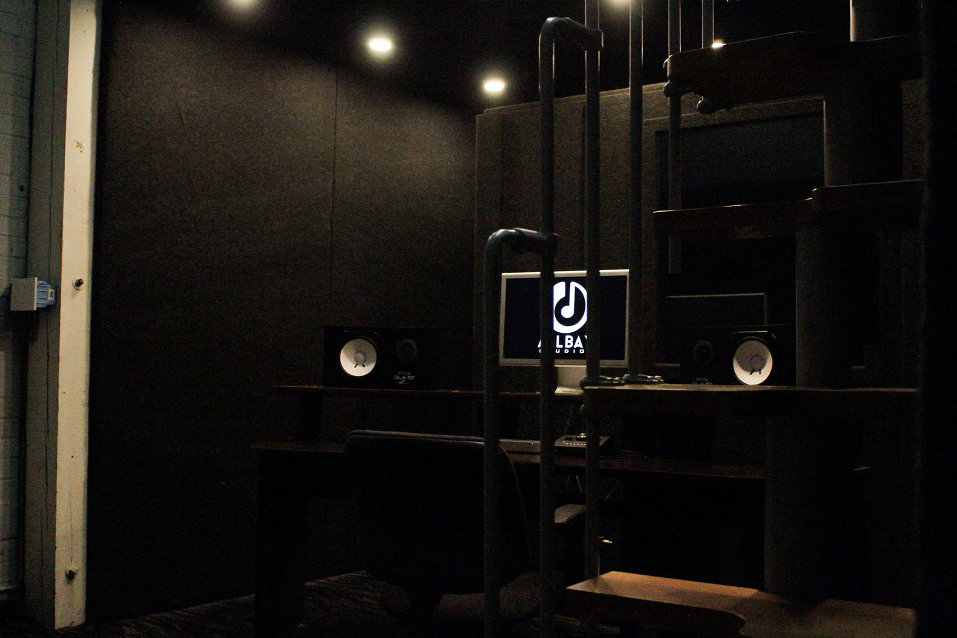 A dark recording studio with a computer on a desk flanked by audio monitors and exposed pipes.