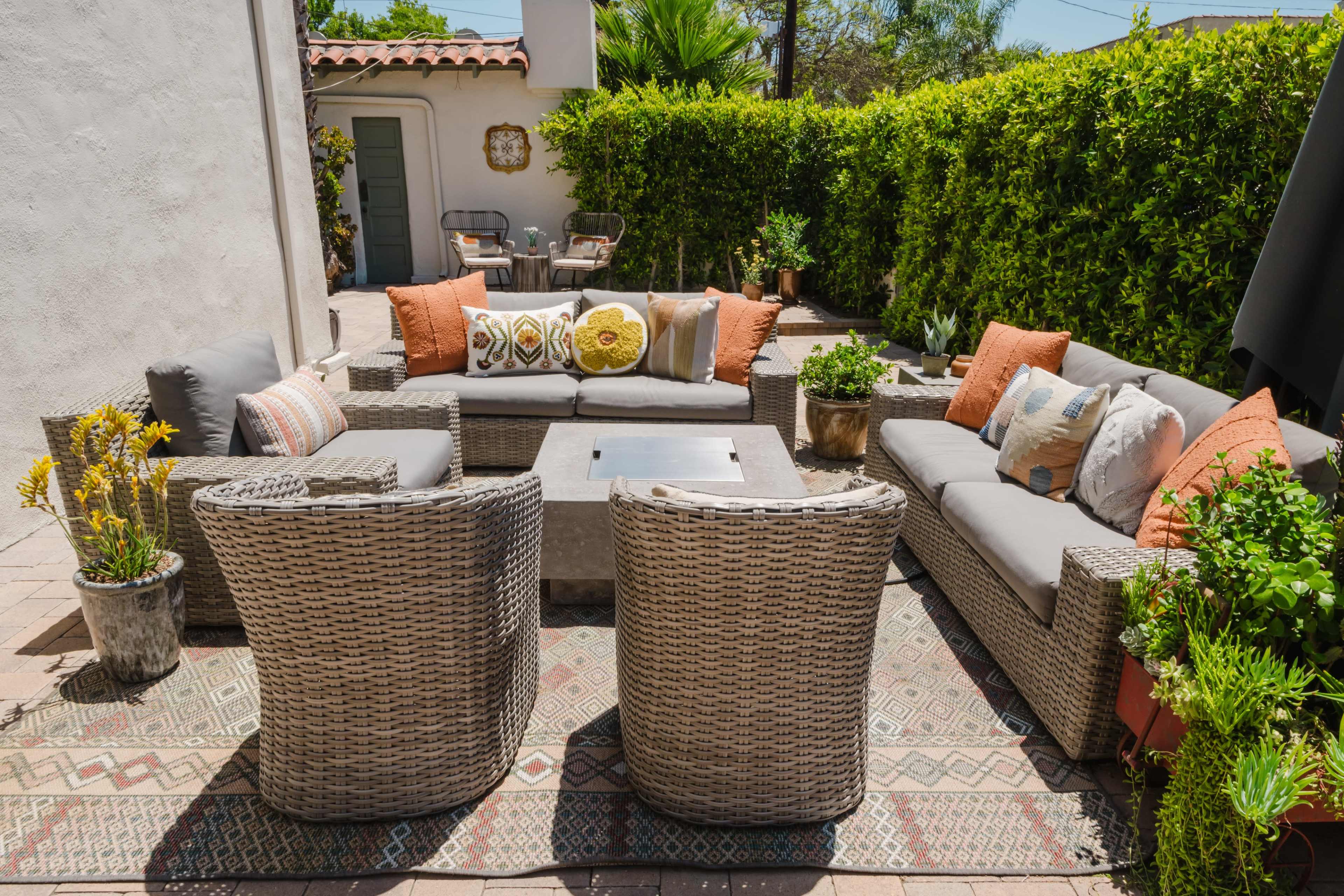 The image shows a patio area featuring woven furniture with cushions, a central fire pit, and various potted plants surrounded by a hedge.