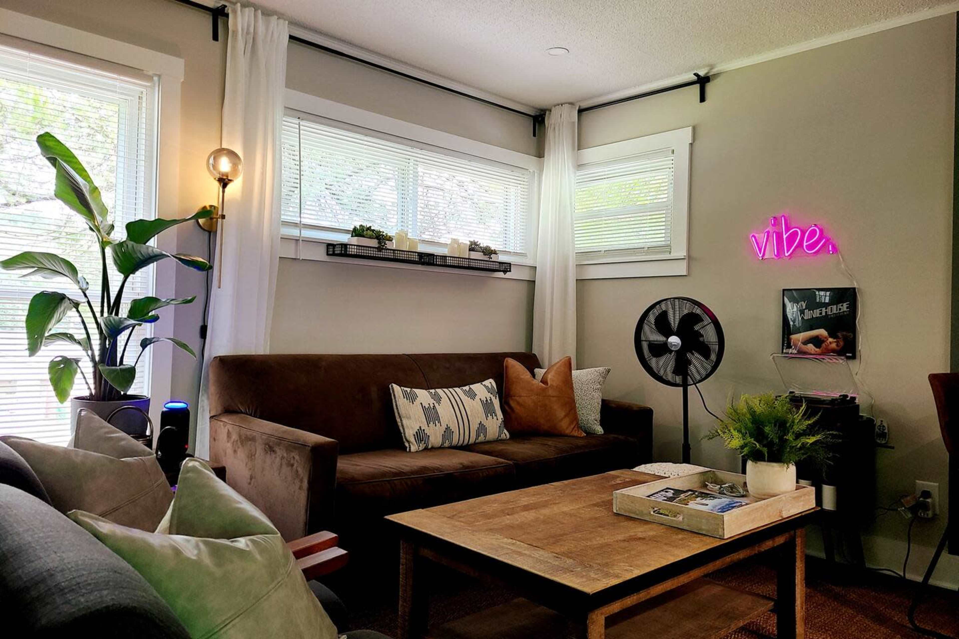 A cozy living room with a brown sofa, a wooden coffee table, and a neon sign that says "vibe," complemented by plants and natural light from the windows.