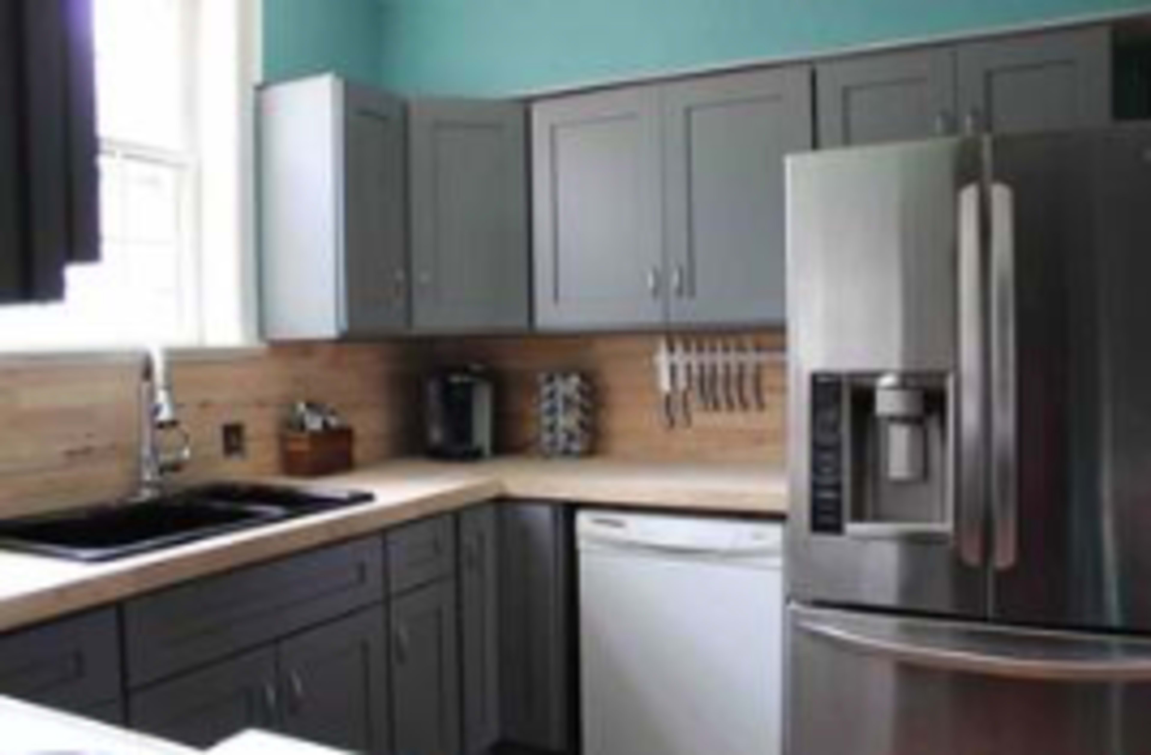 A kitchen with gray cabinetry, a stainless steel refrigerator, a sink, and a dishwasher, all featuring a wooden countertop and a light blue wall.