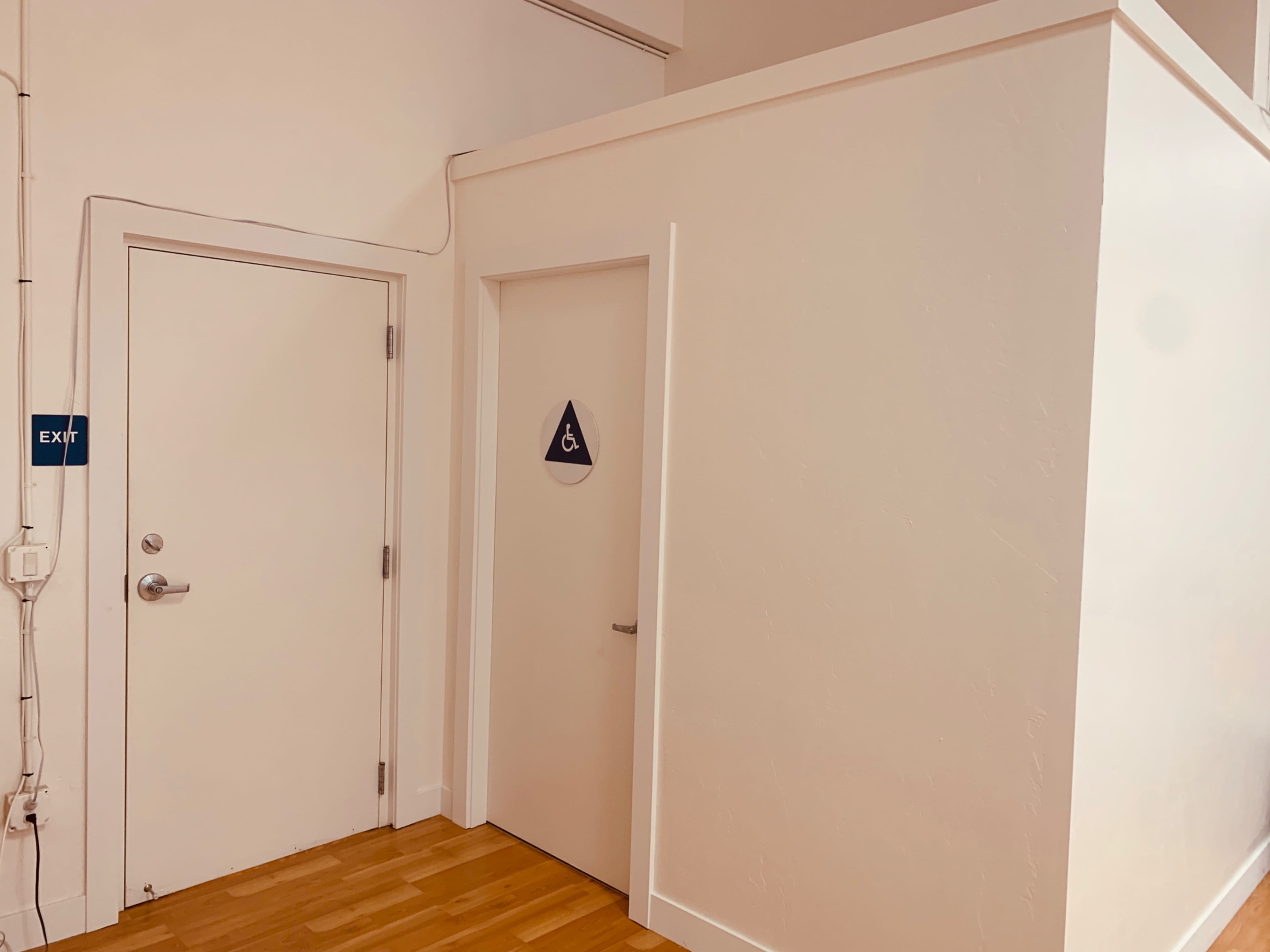 A corner of a room with a white wall, a closed door marked with a restroom symbol, and an exit sign on the adjacent wall.