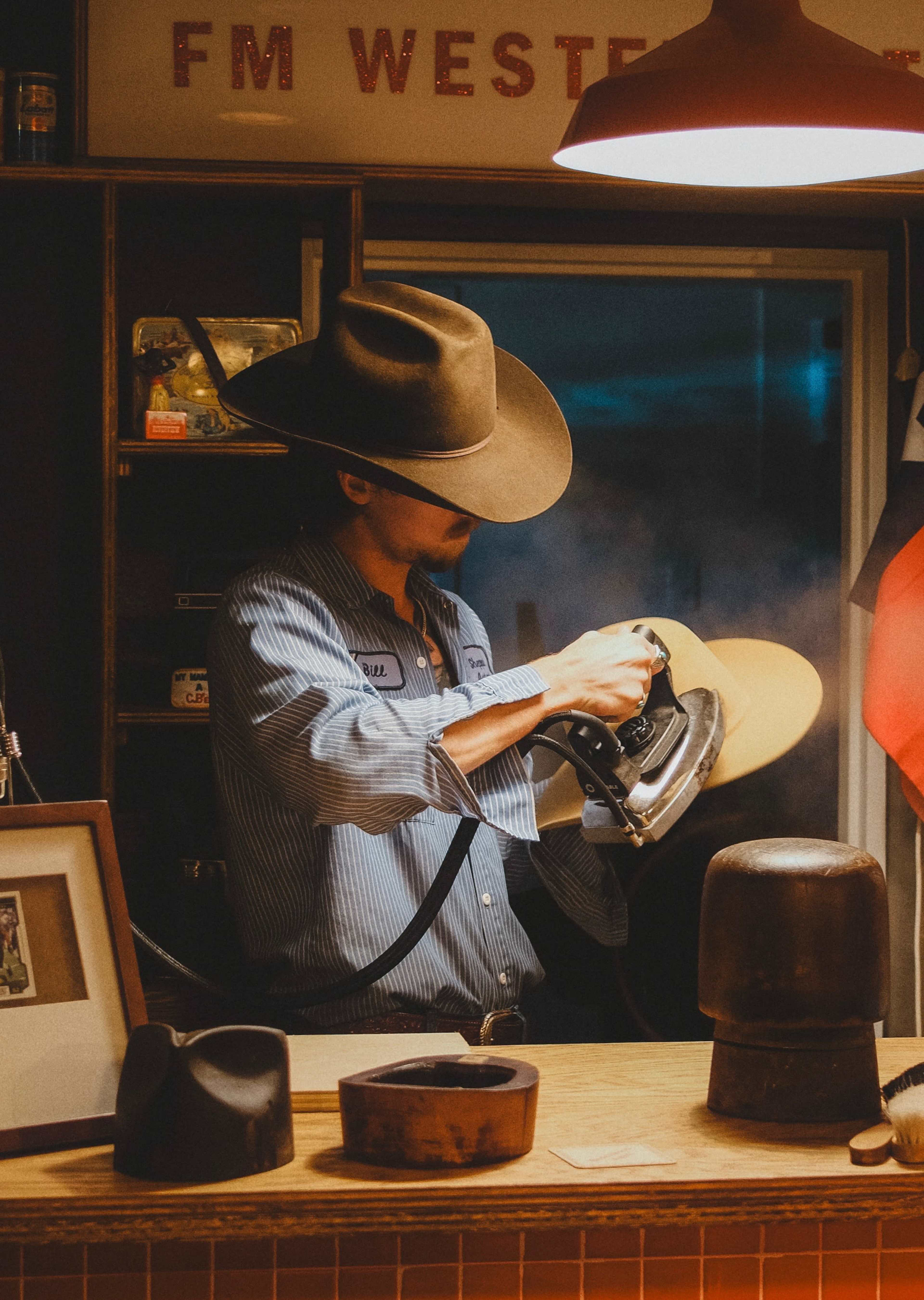 A man wearing a large hat is smoothing a piece of fabric with an iron in a western-themed shop, surrounded by various hats and decor.