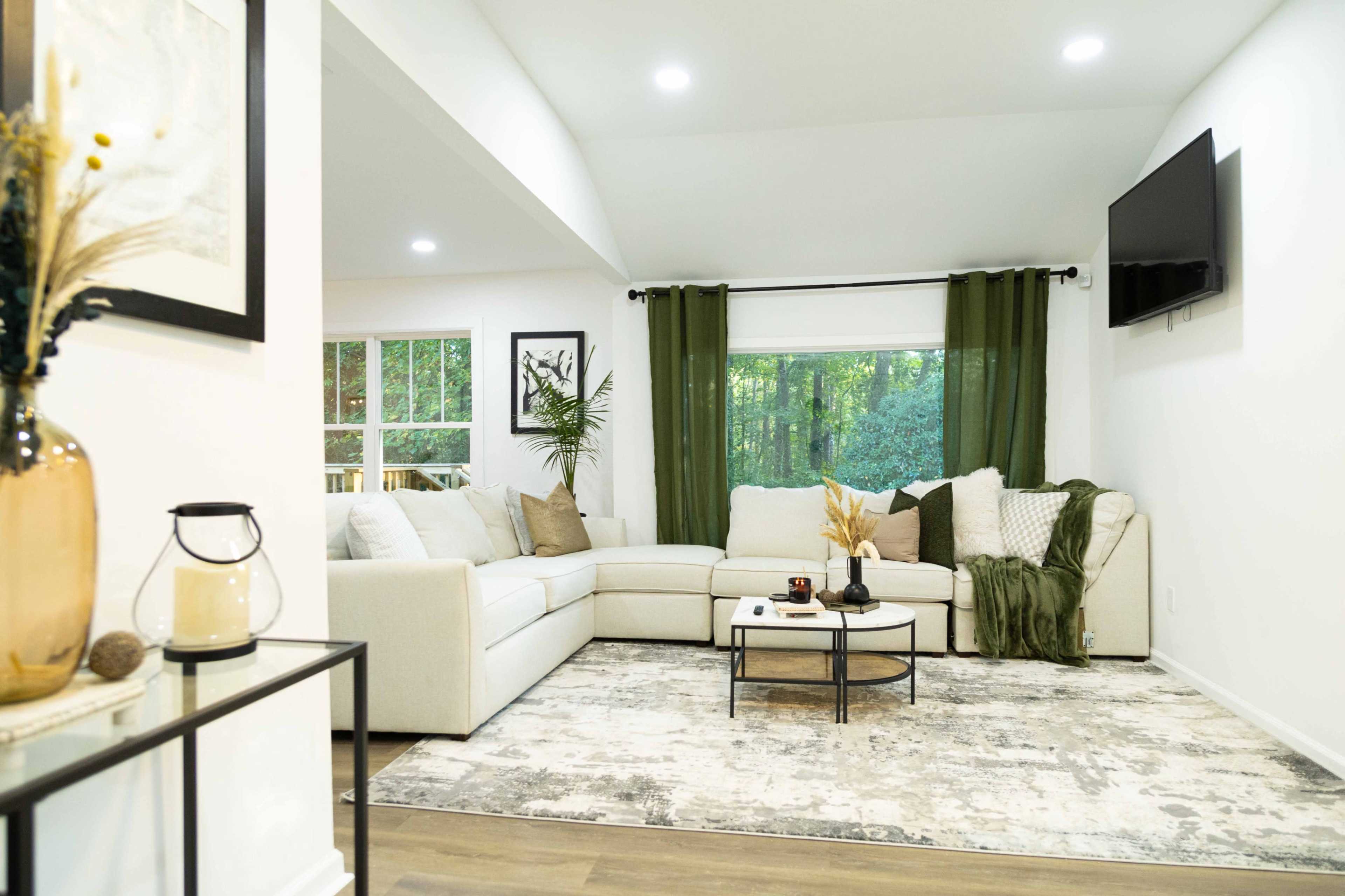 The image shows a modern living room featuring a light-colored sectional sofa, a round coffee table, and large windows with green curtains, surrounded by decorative plants.