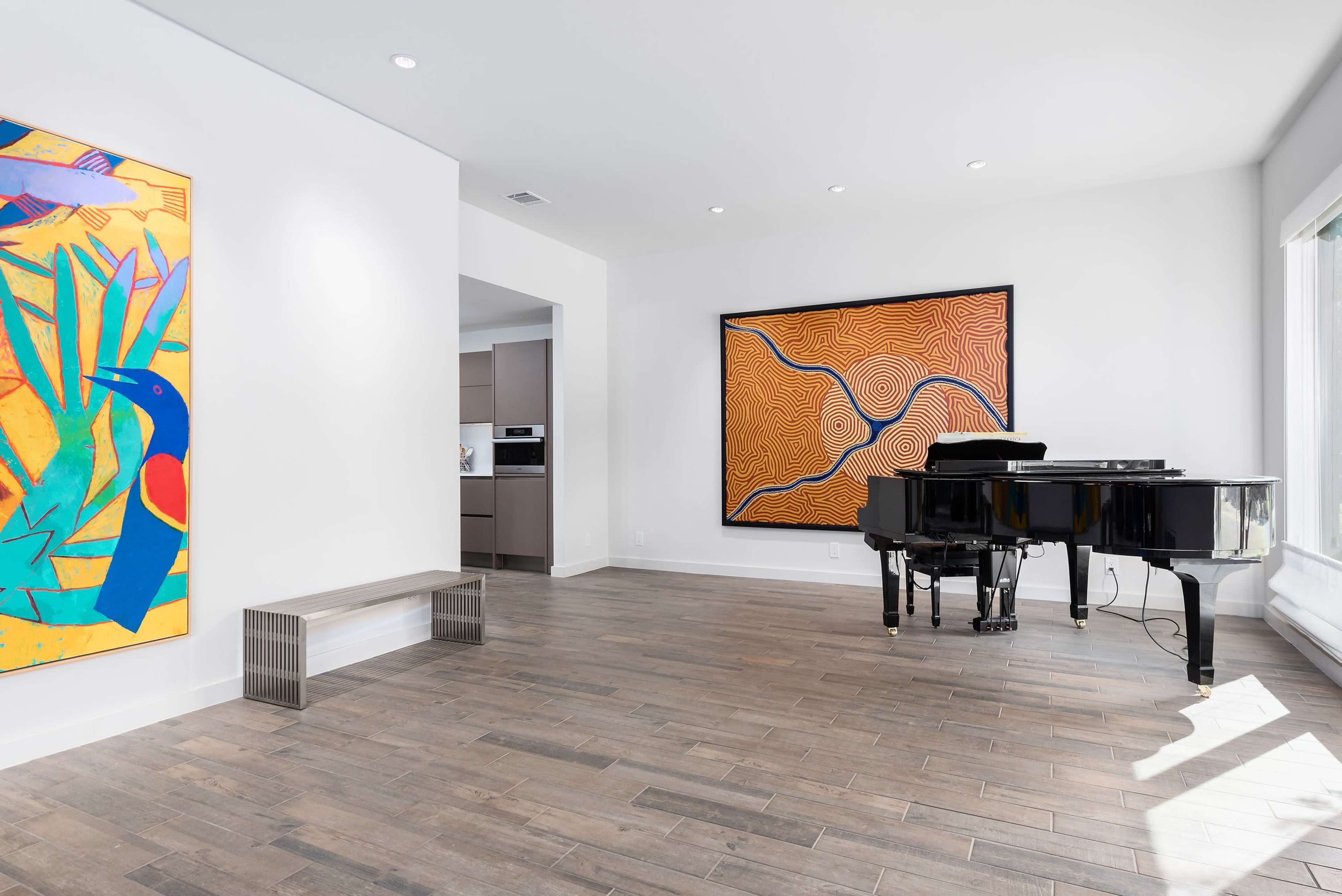A spacious room with a grand piano, two large colorful paintings on the walls, and wood flooring.