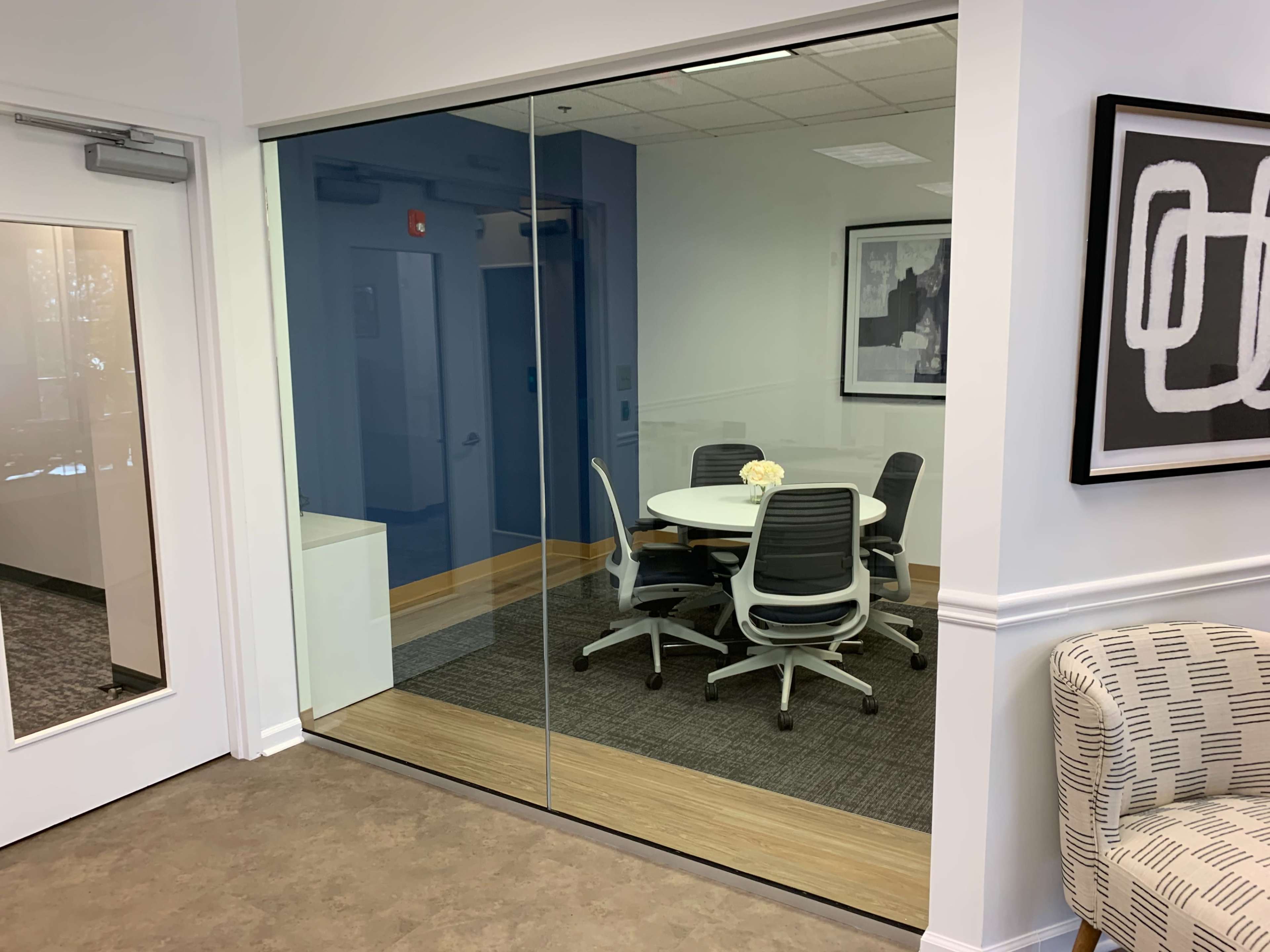 A modern conference room with a round table and five ergonomic chairs is visible through a large glass wall.