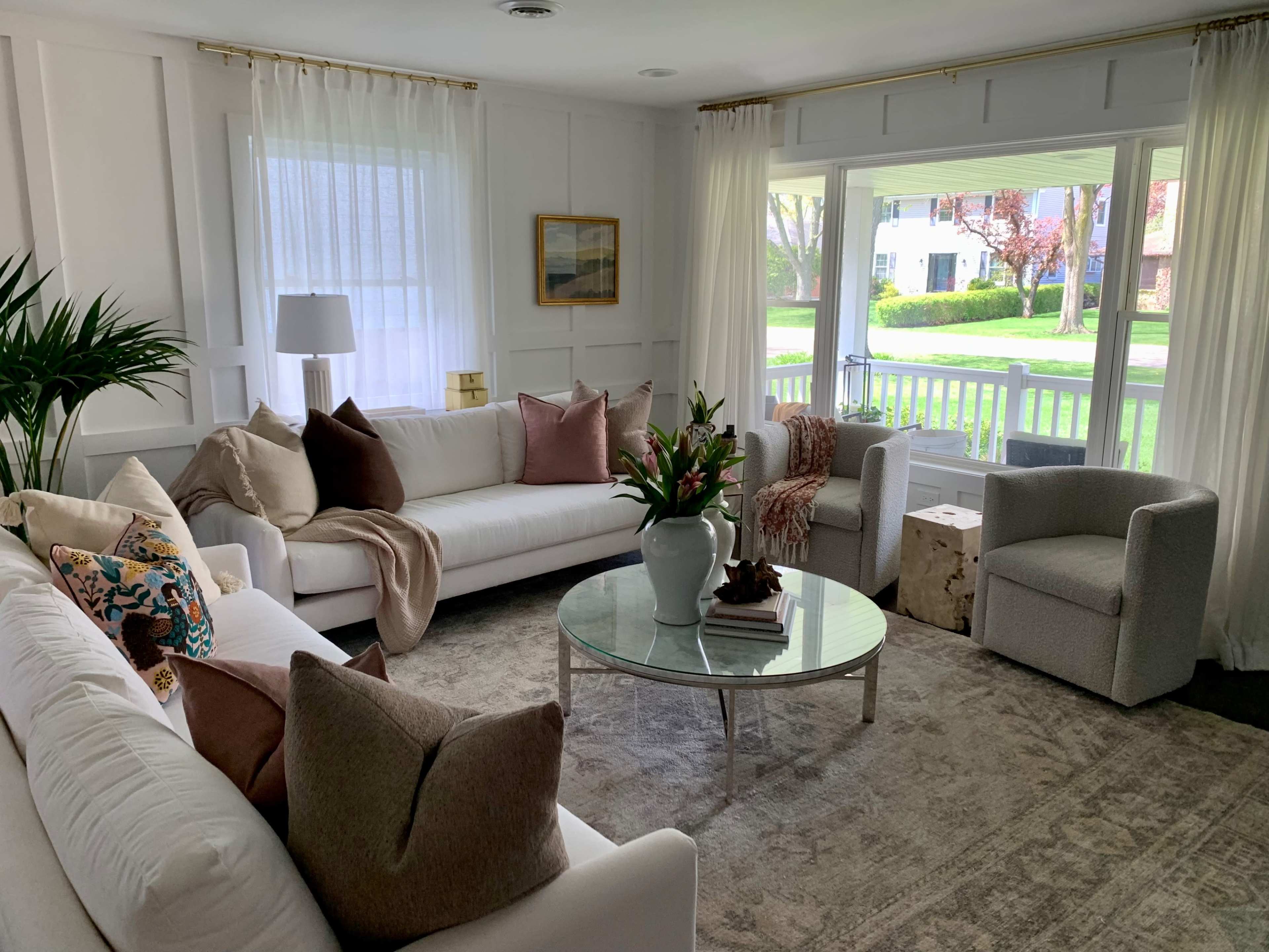 A bright living room featuring a white sectional sofa, a round glass coffee table, and large windows overlooking a green yard with trees.