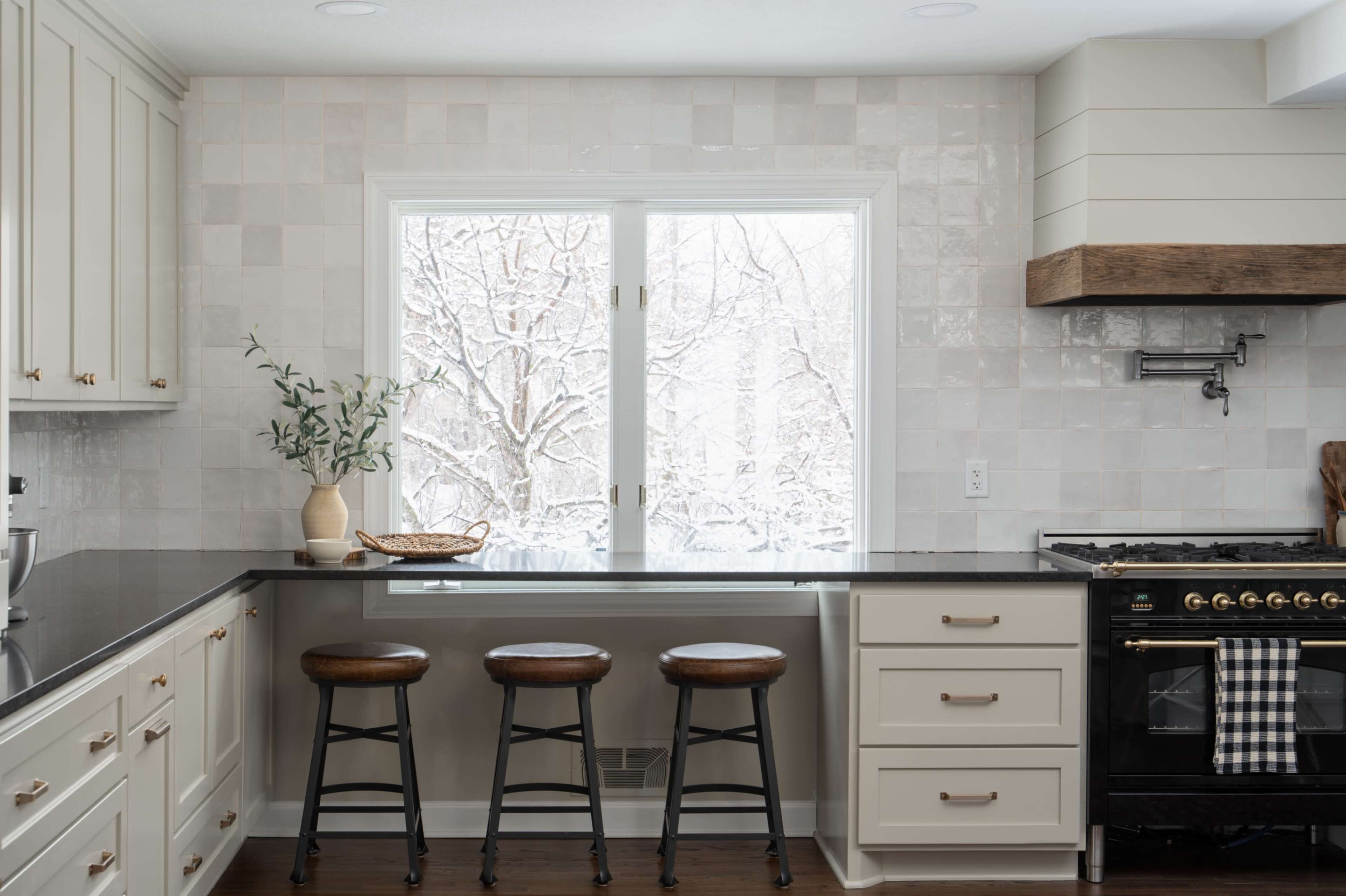 A modern kitchen features three wooden stools at a countertop with a large window overlooking a snowy landscape.