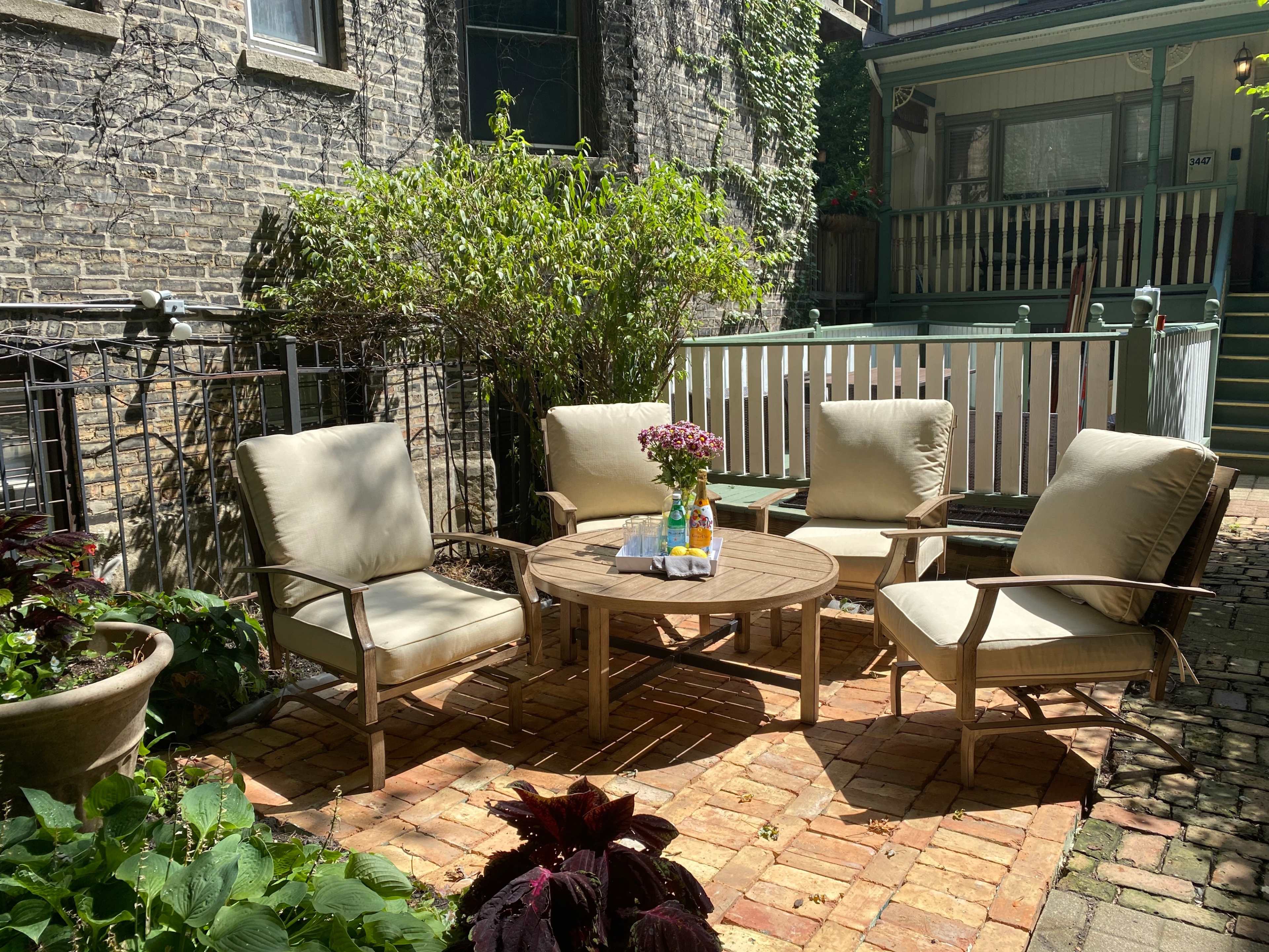 A patio area features four cushioned chairs around a round wooden table, with a vase of flowers on top, surrounded by greenery and brick flooring.