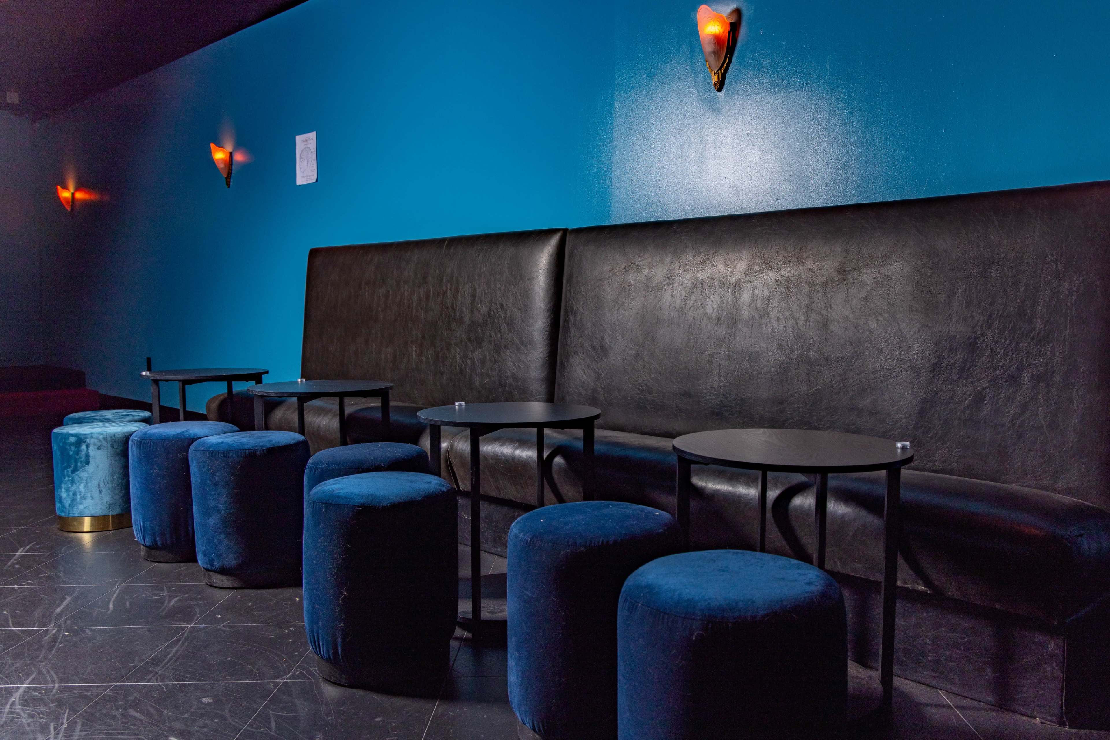A dark leather bench is against a blue wall, accompanied by several small black tables and blue upholstered stools.