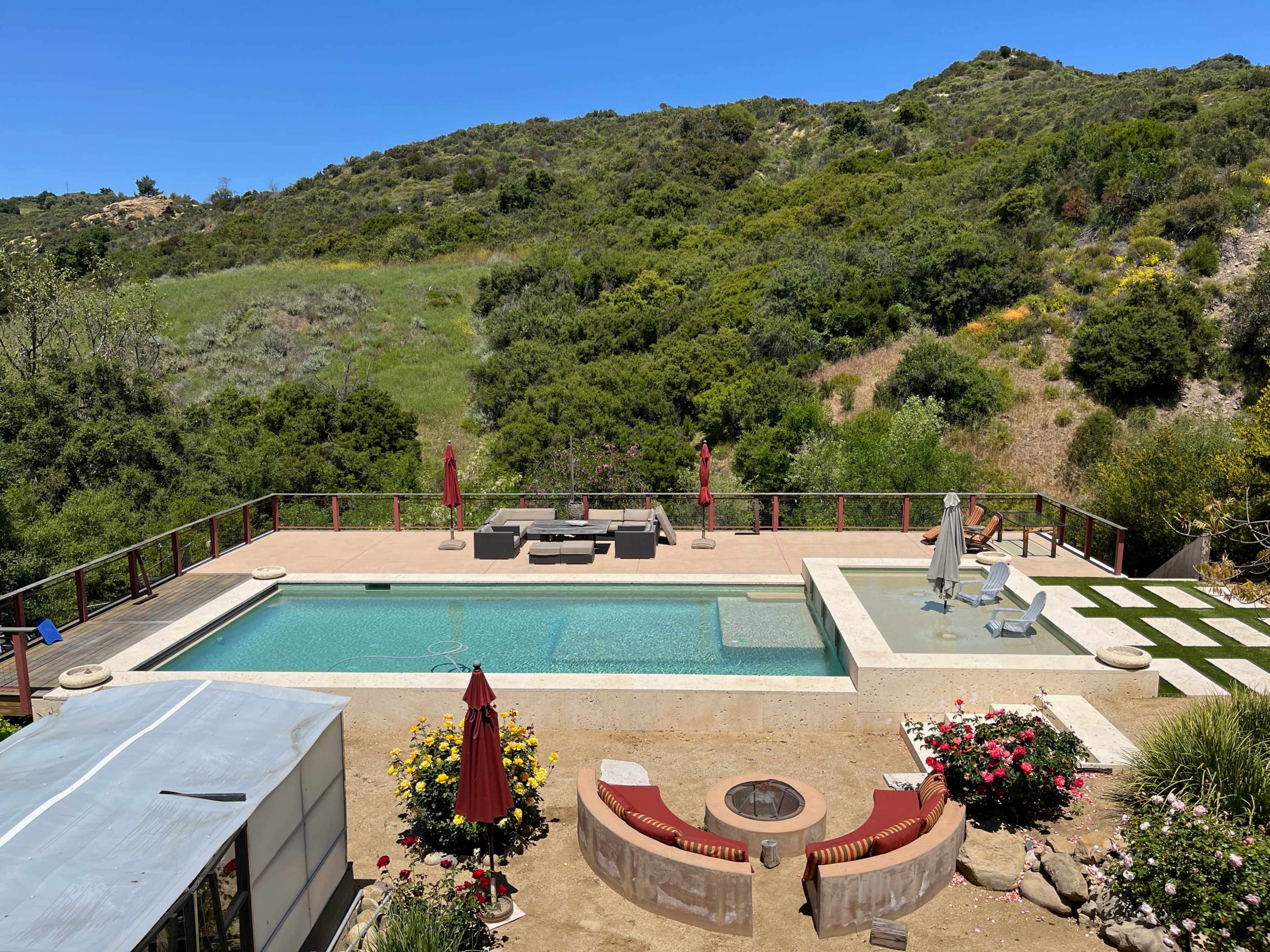 The image shows a backyard with a swimming pool surrounded by a deck, lounge chairs, and a seating area, set against a backdrop of lush green hills.