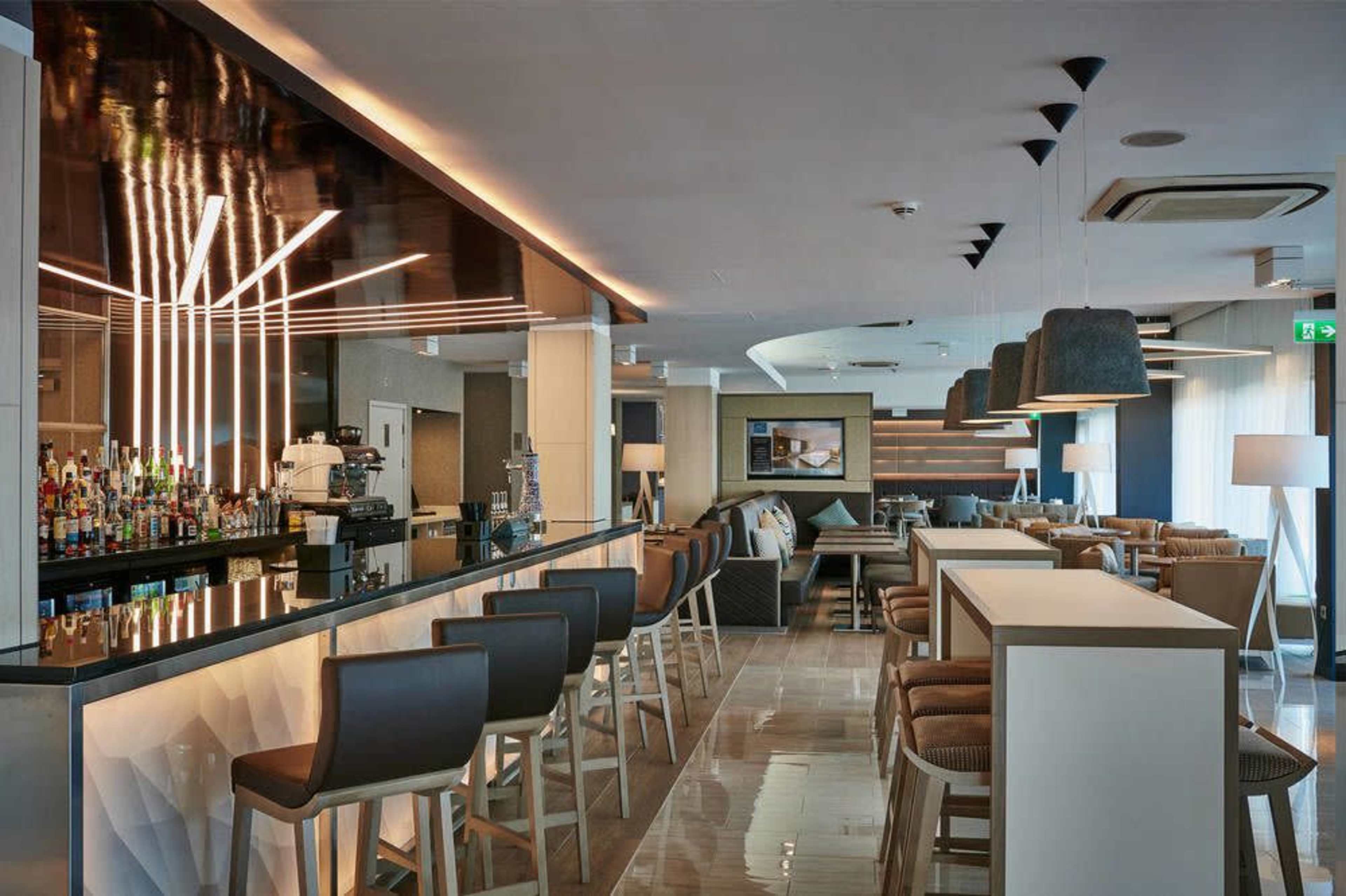 A modern bar area with a sleek counter, high stools, and a variety of bottles displayed on shelves.