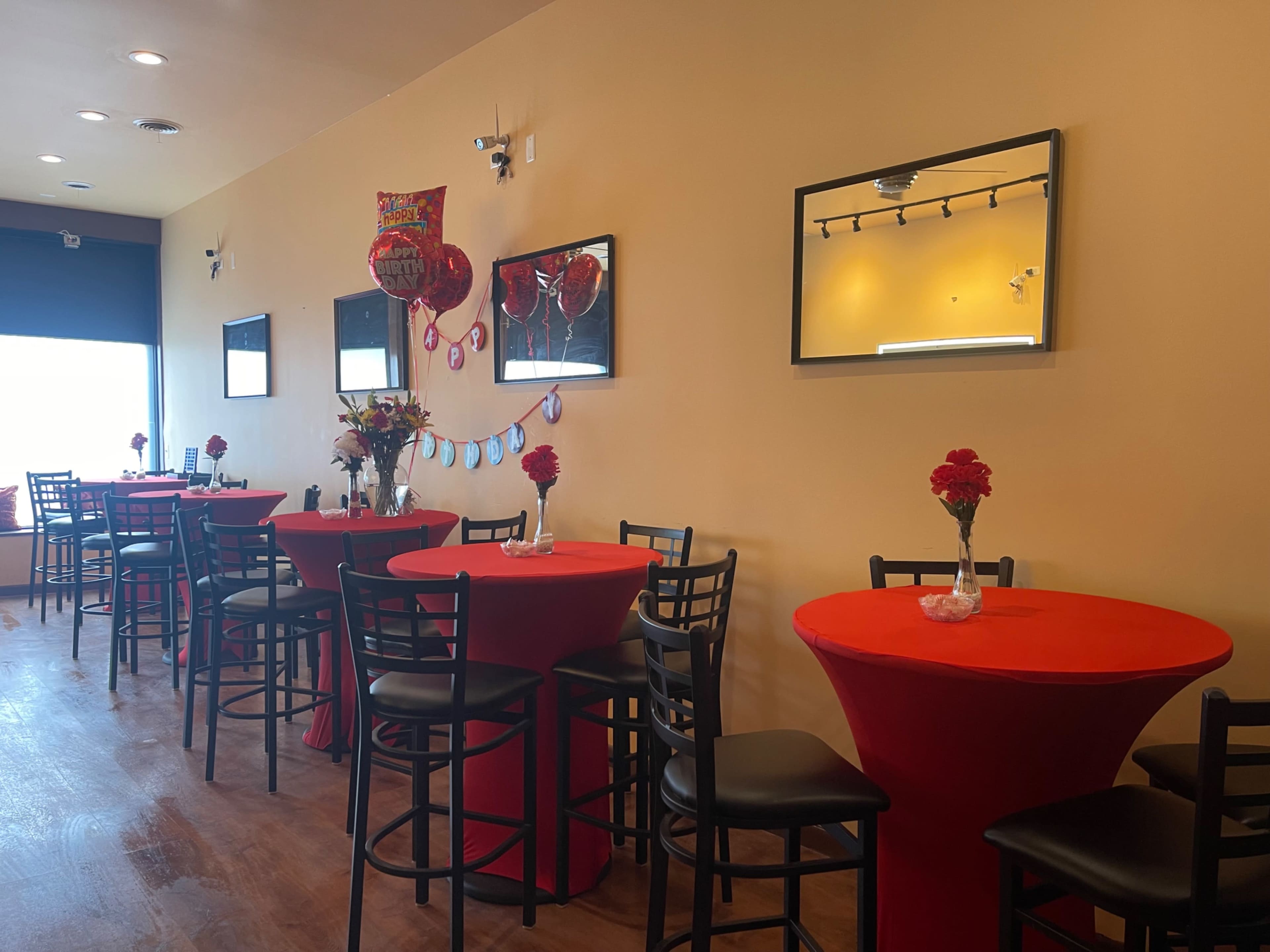 A brightly lit restaurant interior featuring tables with red tablecloths, black chairs, and wall decorations, including mirrors and framed artwork.