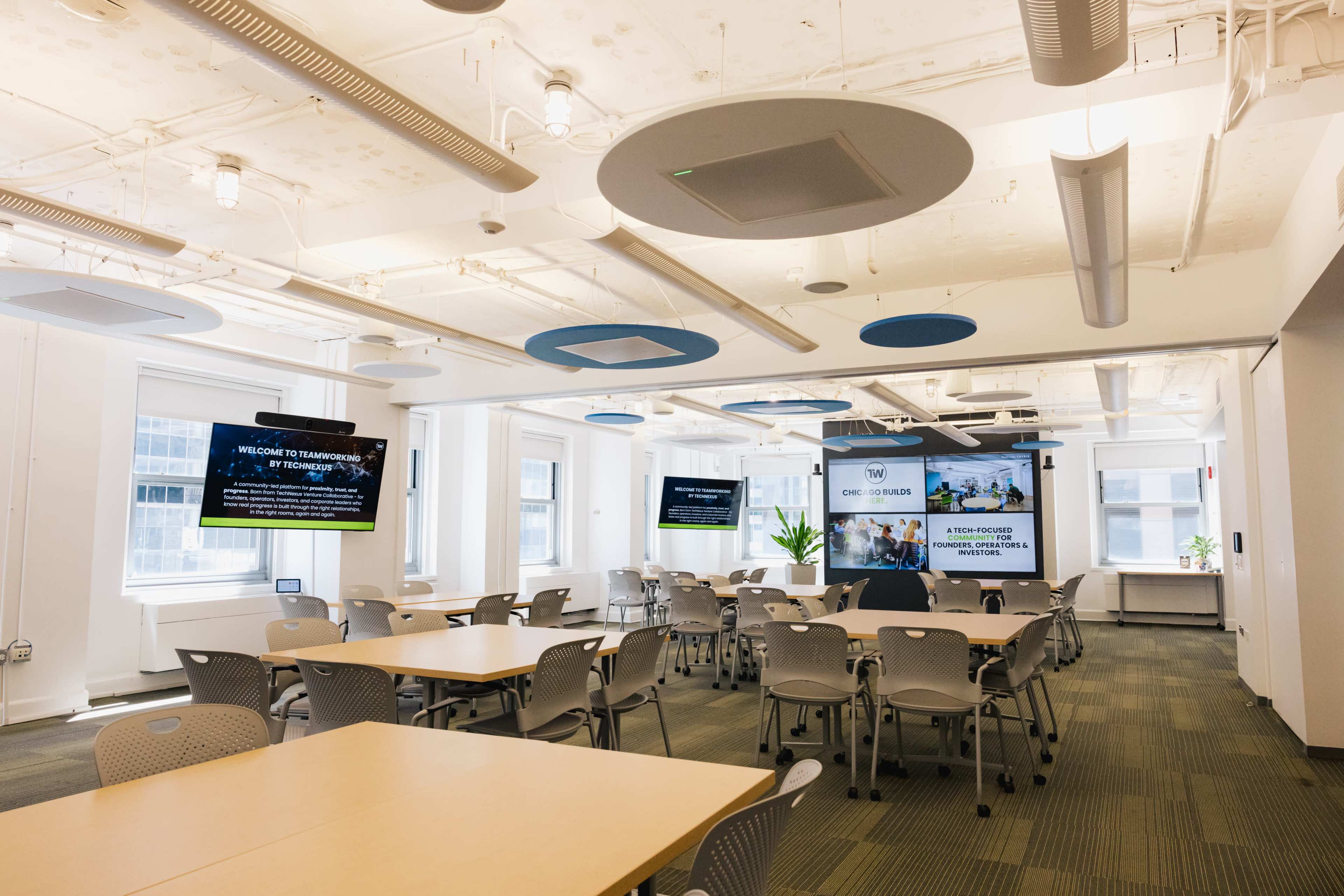 The image shows a modern conference room with multiple tables and chairs, equipped with large screens displaying presentations and ample natural light.