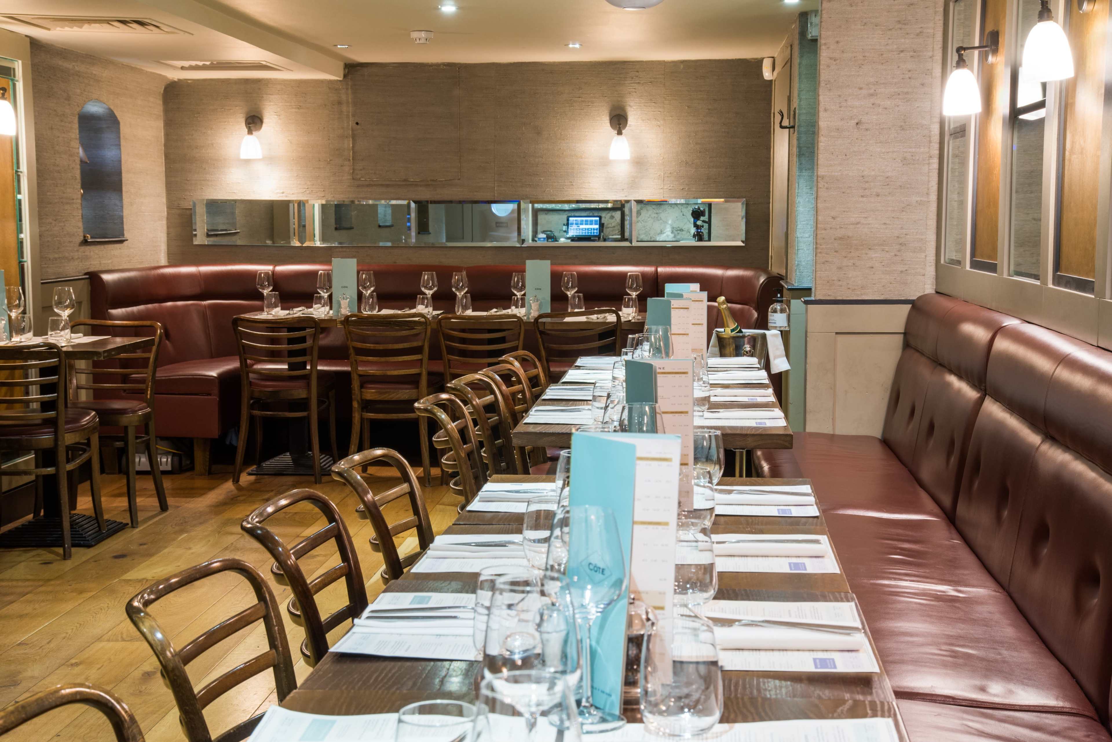 The image shows a restaurant interior with neatly arranged tables and chairs, set with menus and glassware, under warm lighting.