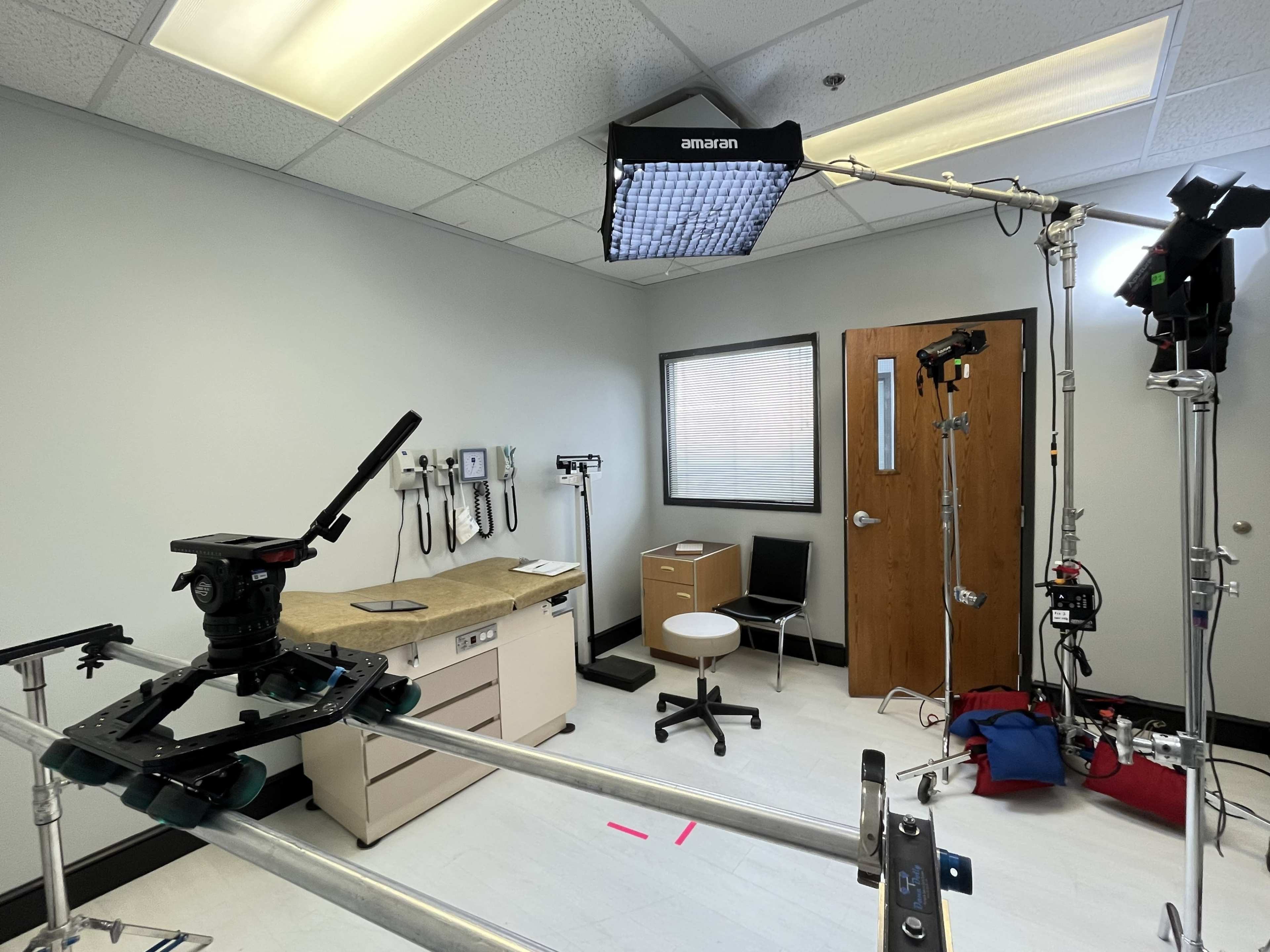 The image shows a medical examination room with a camera setup, lighting equipment, and a stool next to a doctor's desk.