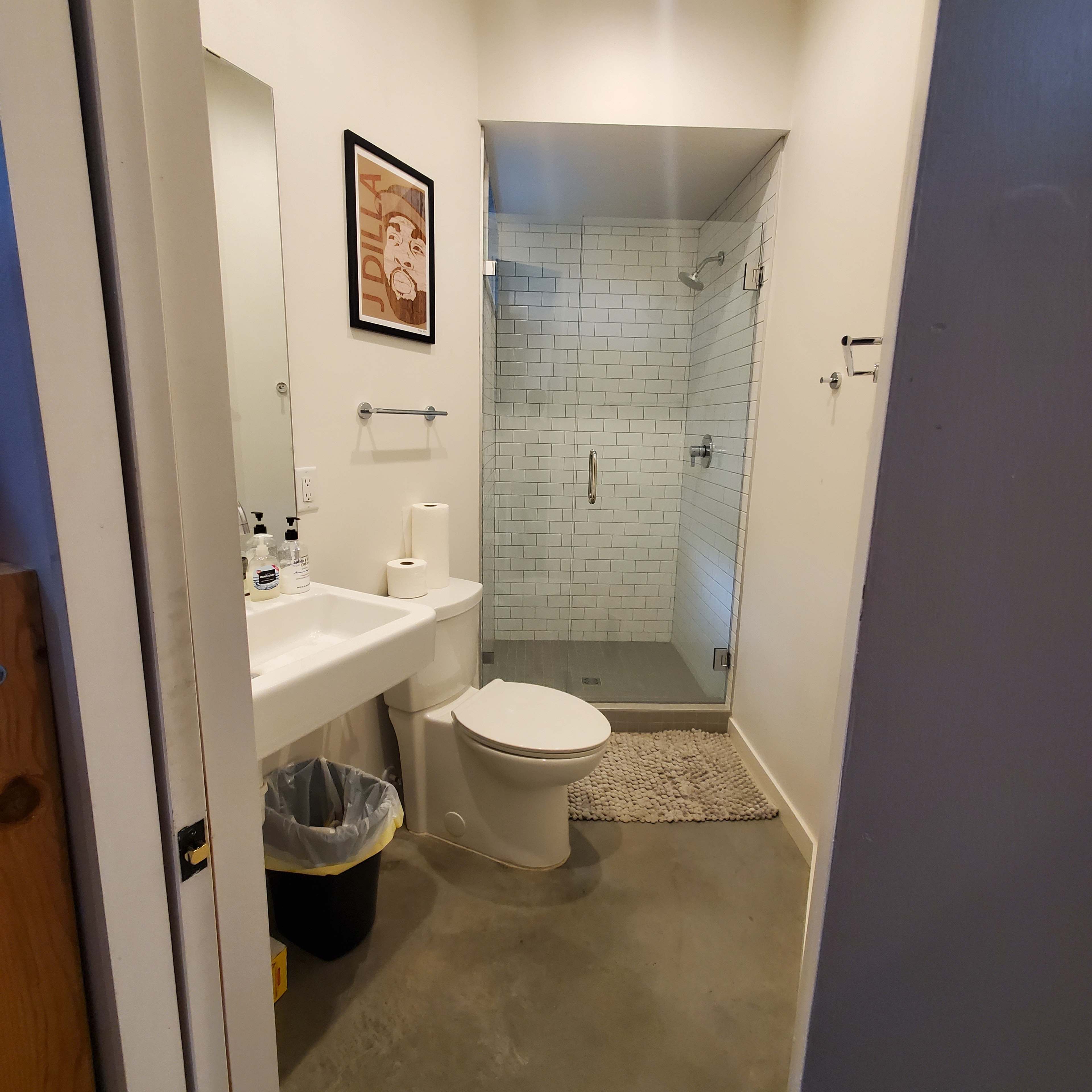 A small bathroom features a shower, toilet, sink, and a trash can, with white tile and concrete flooring.