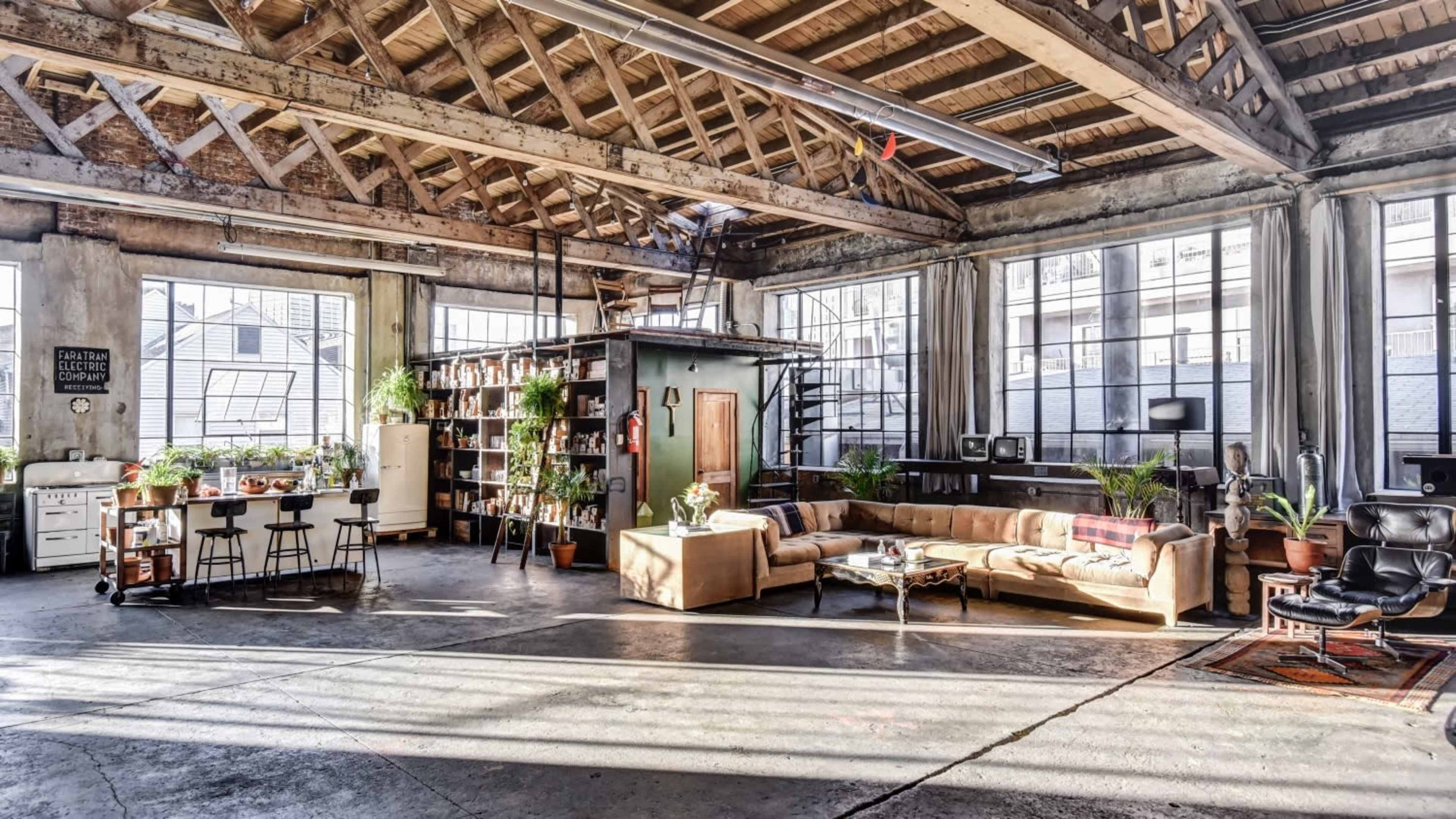 A spacious, industrial-style loft filled with natural light, featuring a seating area, a kitchen space, and various plants throughout.