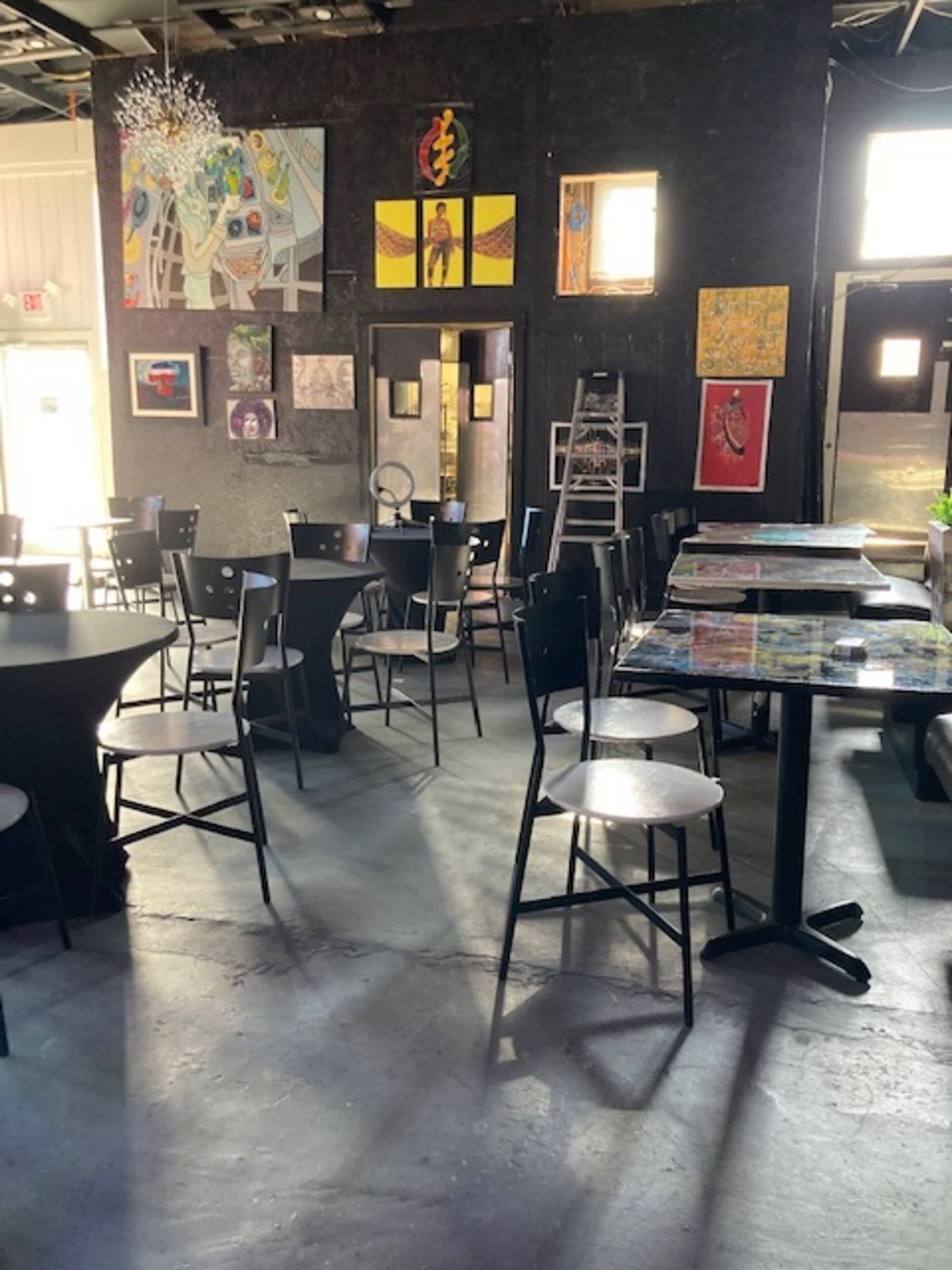 The interior of a café features black tables and chairs, colorful artworks on the walls, and a ladder leaning against the wall.