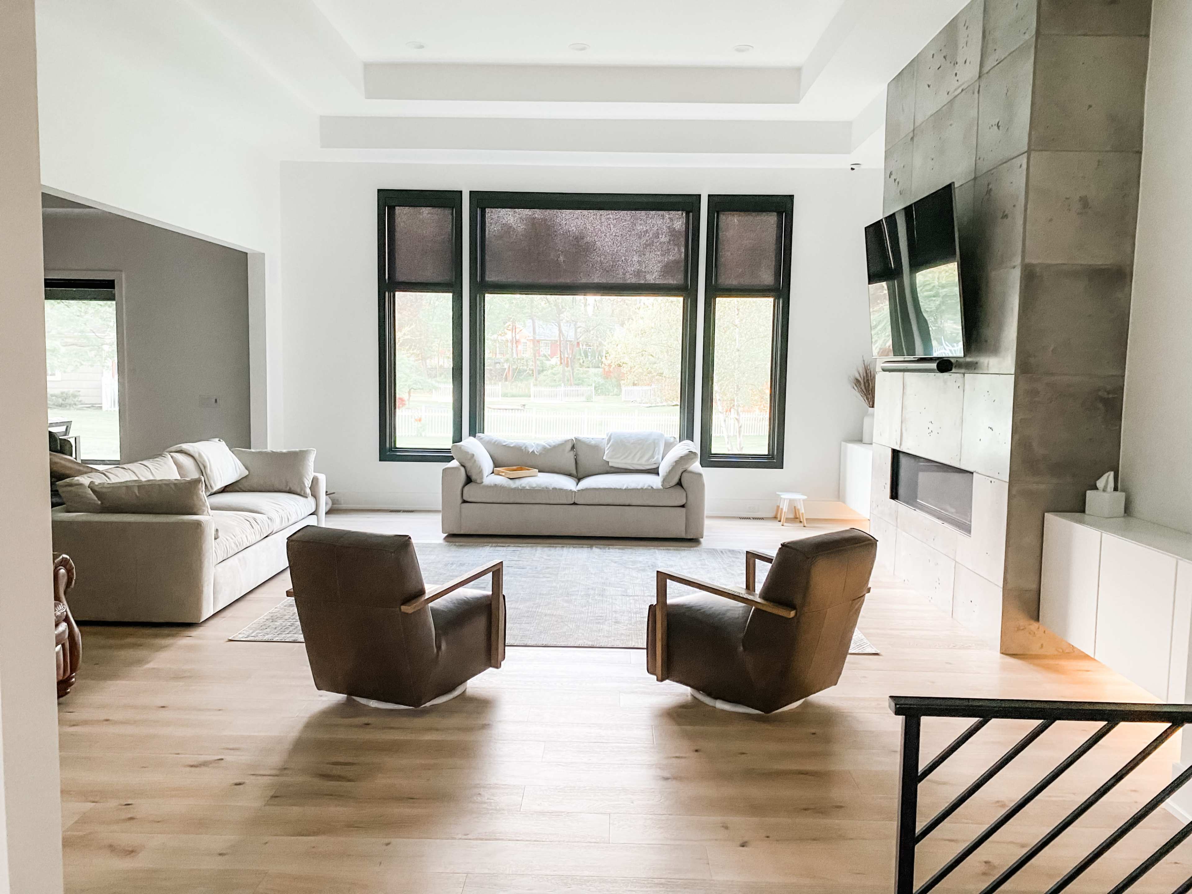 A modern living room features a light-colored sofa, two brown armchairs, large windows overlooking a garden, and a minimalist fireplace.