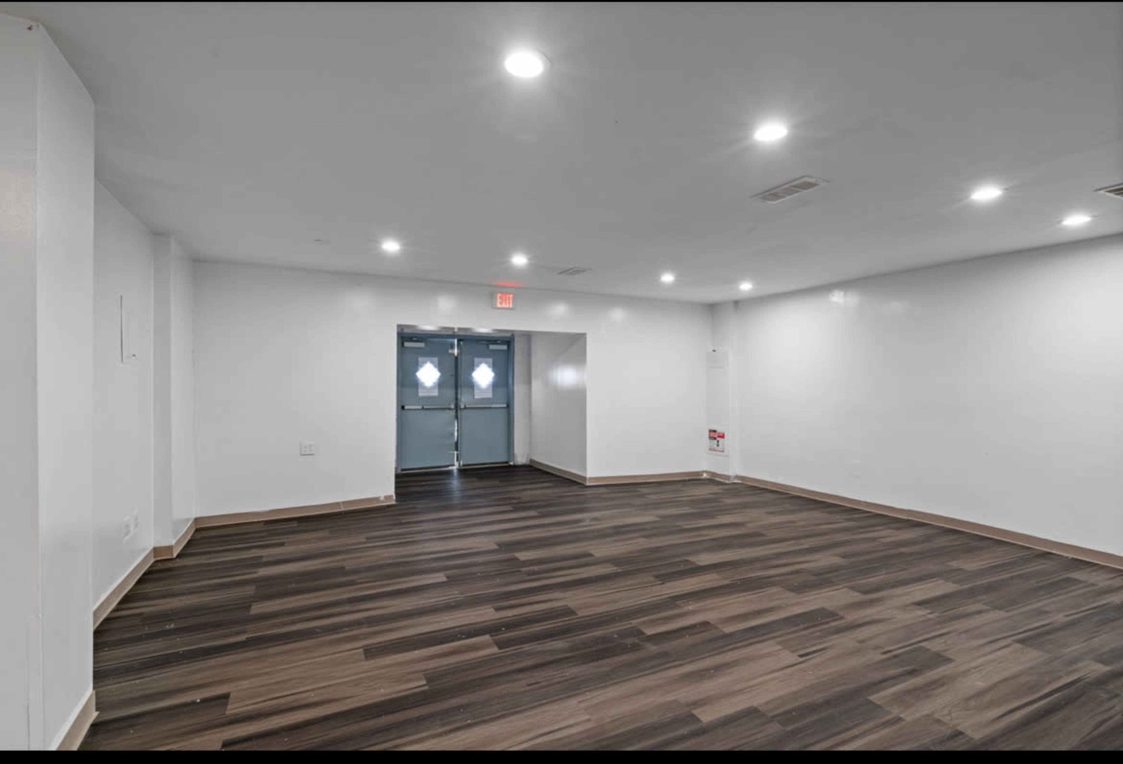 The image shows an empty room with white walls and hardwood flooring, featuring recessed lighting and a set of double doors at one end.