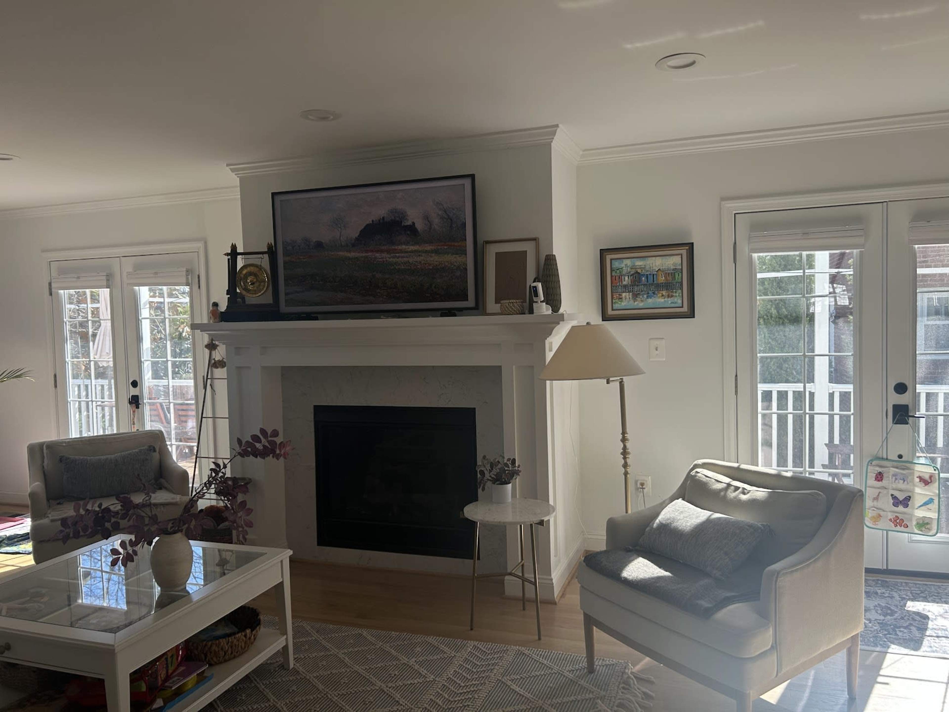 The image shows a well-lit living room featuring a fireplace with a television mounted above it, a comfortable armchair, a decorative table, and large windows leading to a balcony.