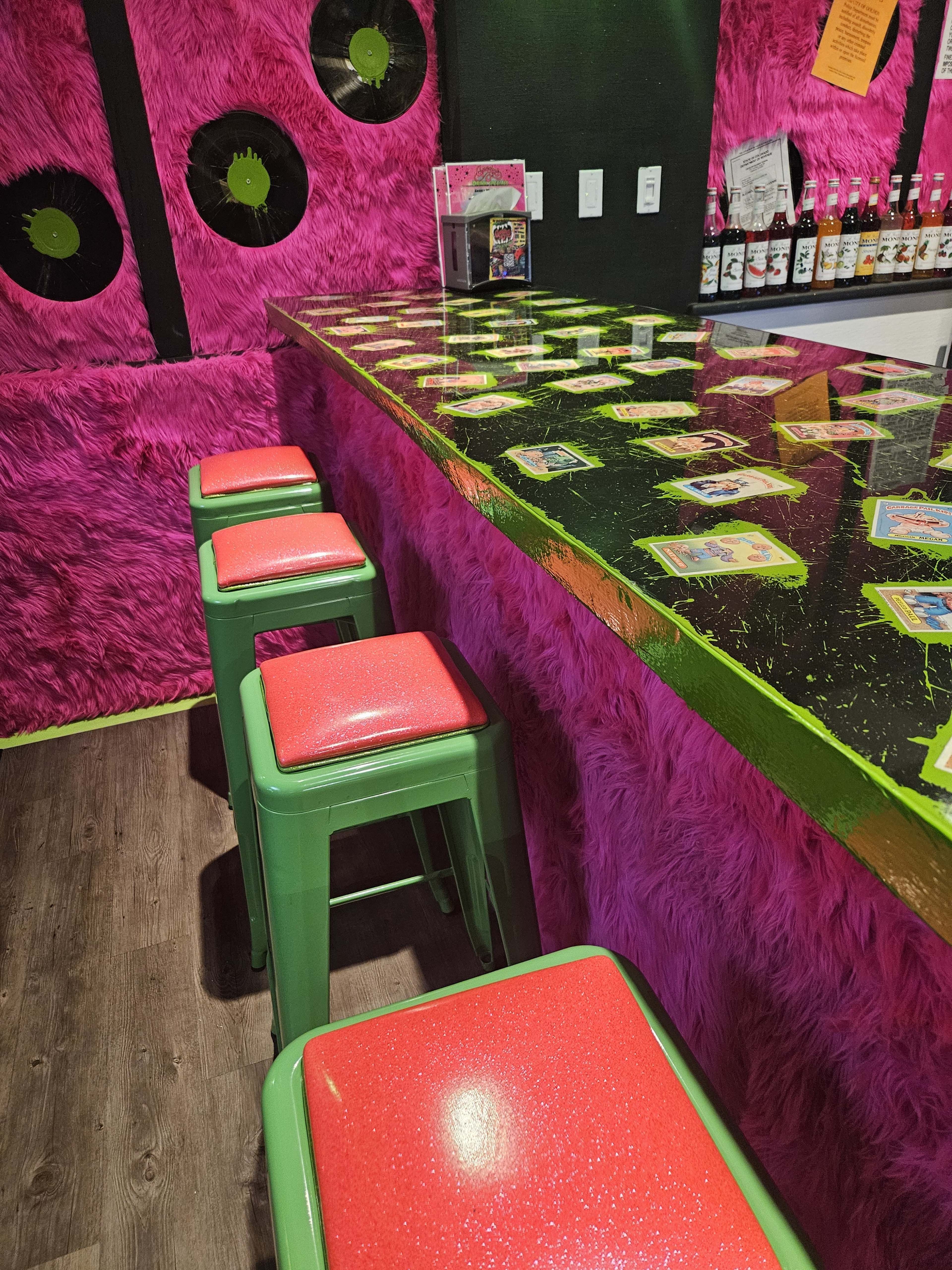 The image shows a brightly colored bar area with pink fur-covered walls, a glossy black countertop, and green stools with red seats.