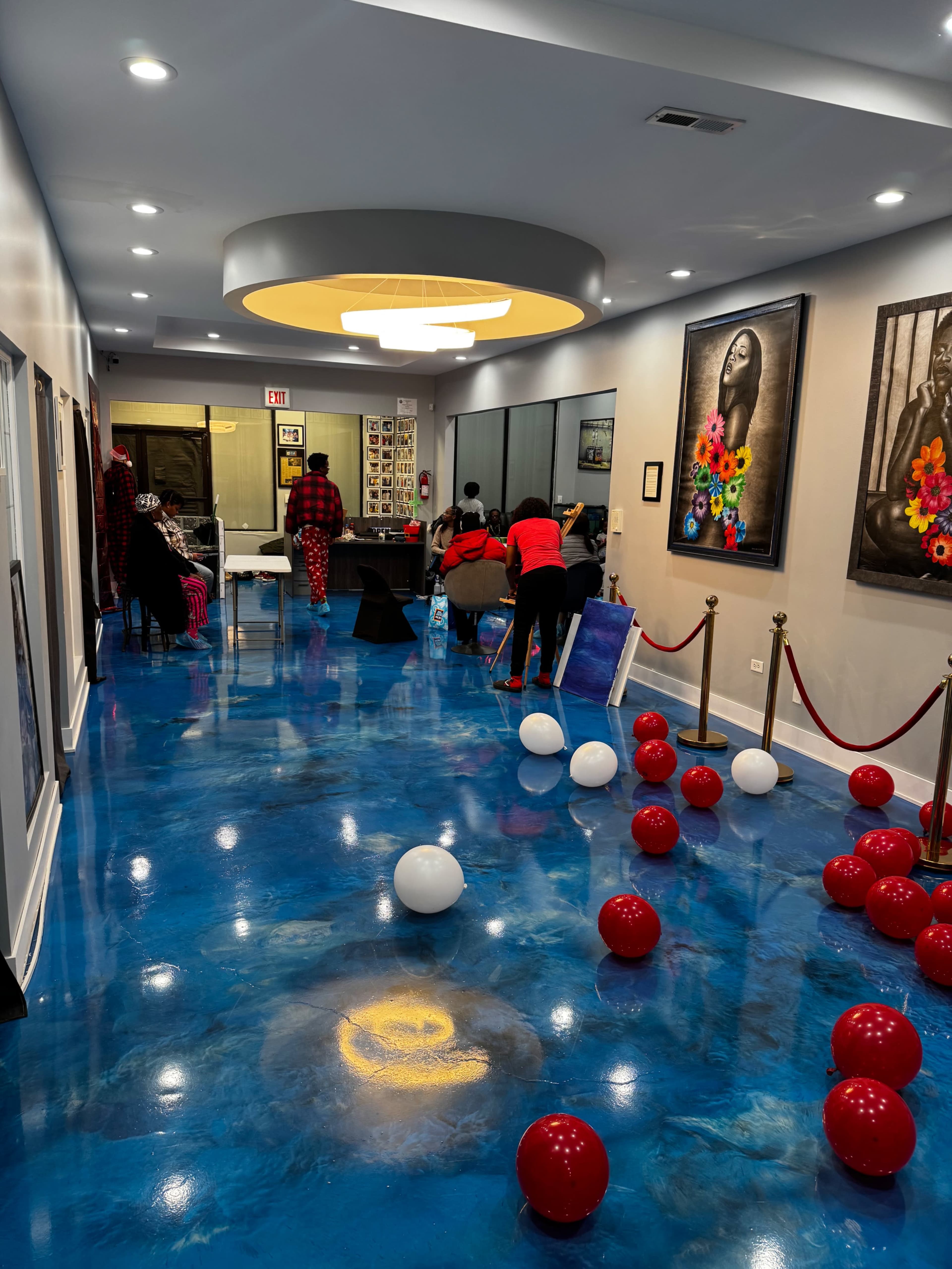 The scene shows a brightly lit hallway decorated with red and white balloons, featuring colorful artwork on the walls and people engaged in activities at a table at the end.