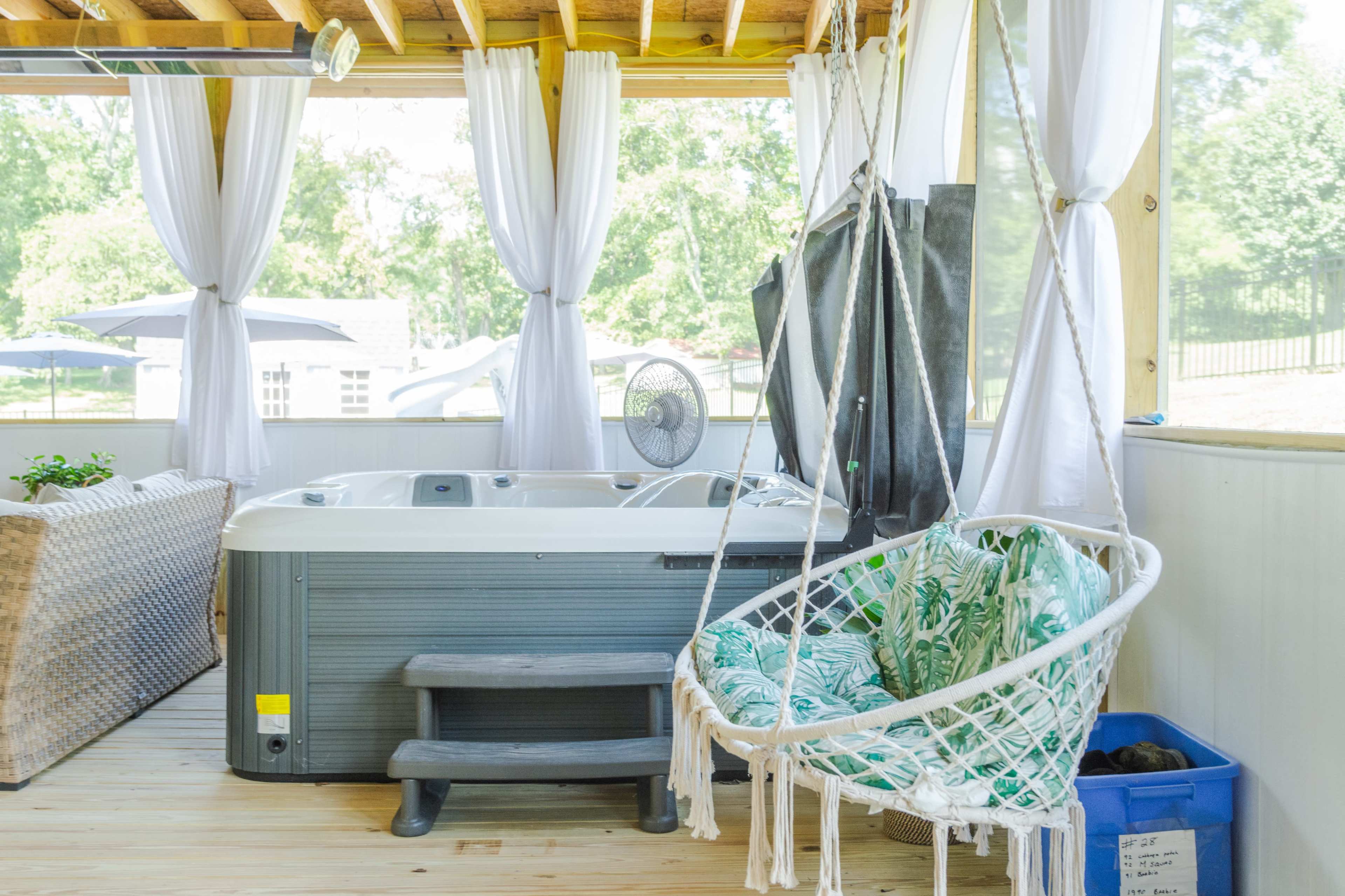 The image shows a spacious indoor area featuring a hot tub beside a hanging chair with a tropical pattern, large windows allowing natural light, and white curtains.