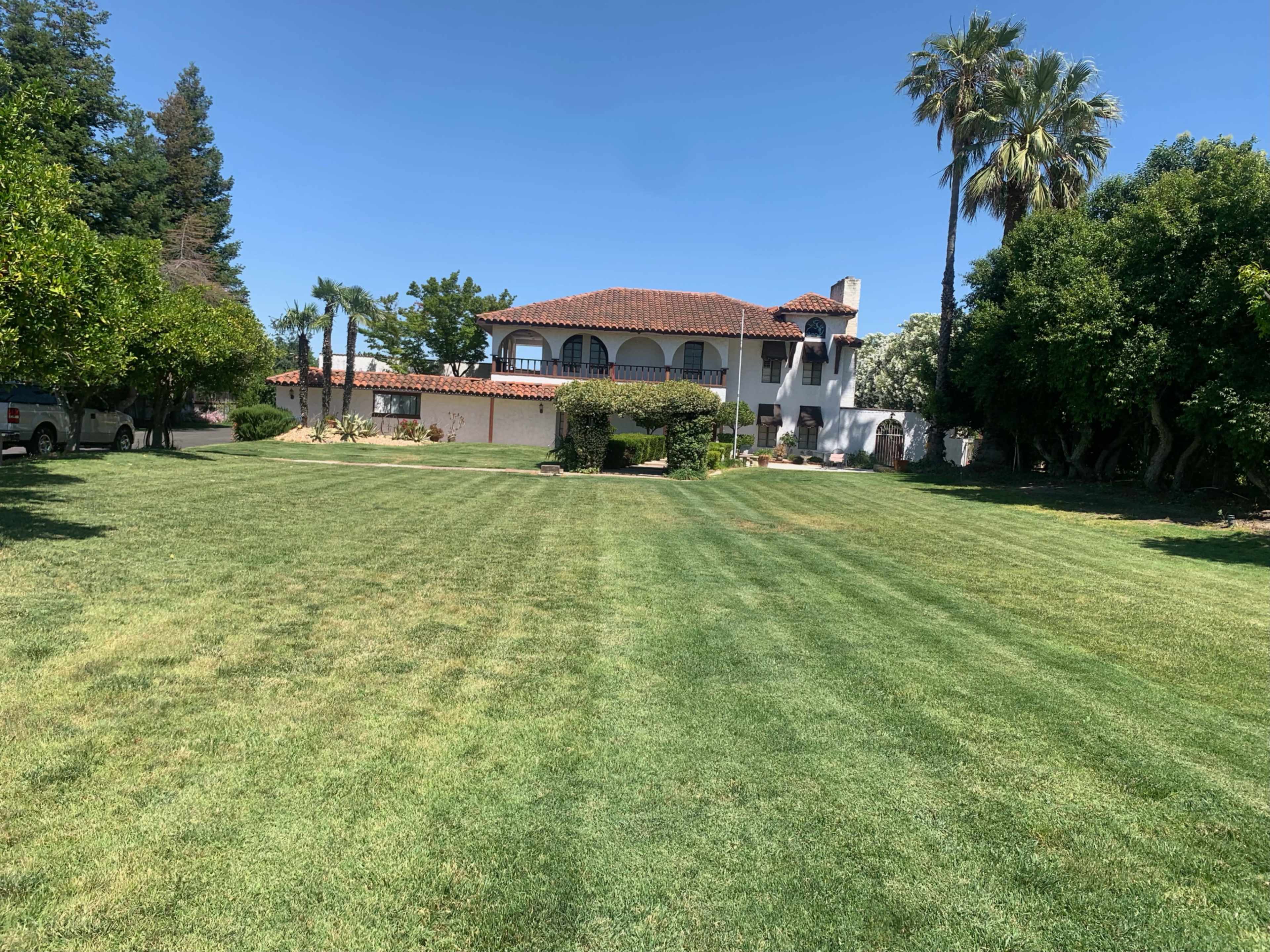 A large, two-story house with a tile roof is situated behind a neatly manicured lawn and surrounded by palm trees and various shrubs.