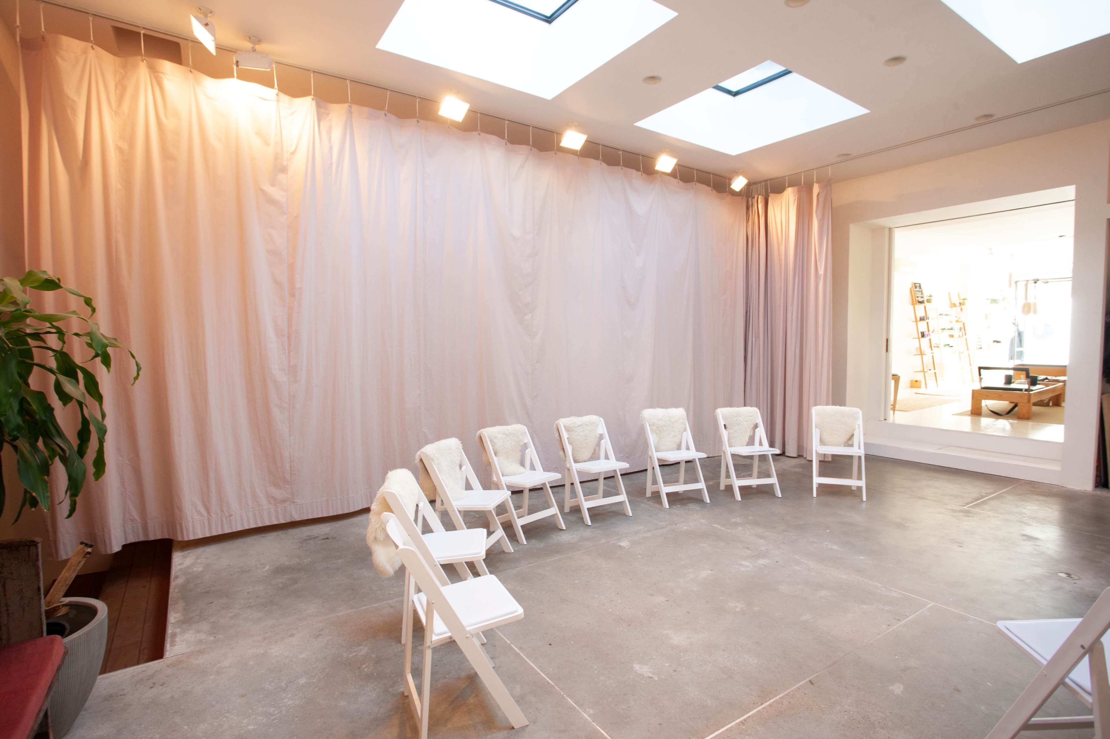 The image shows a circular arrangement of white chairs in a well-lit room with a curtain backdrop and skylights.