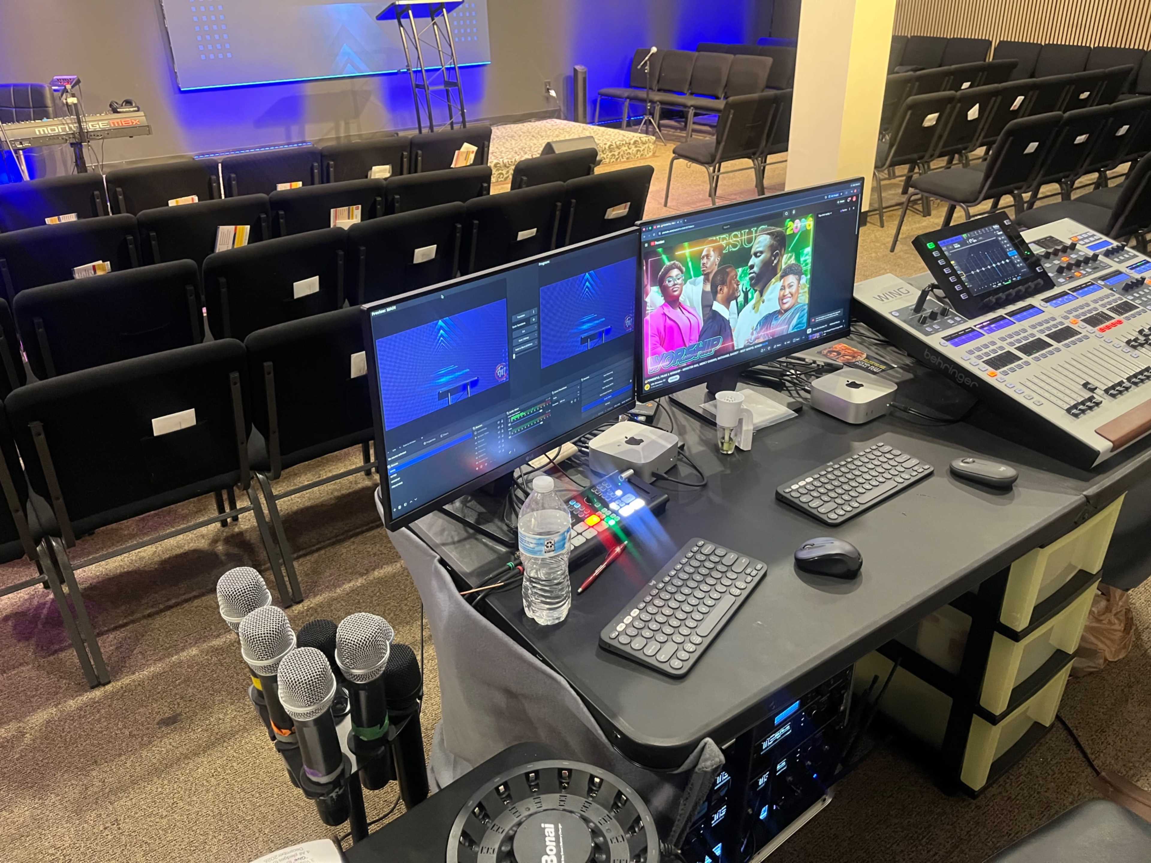 The image shows a control station with two computer monitors, a sound mixing console, microphones, and a water bottle, positioned in front of an empty seating area in a venue.