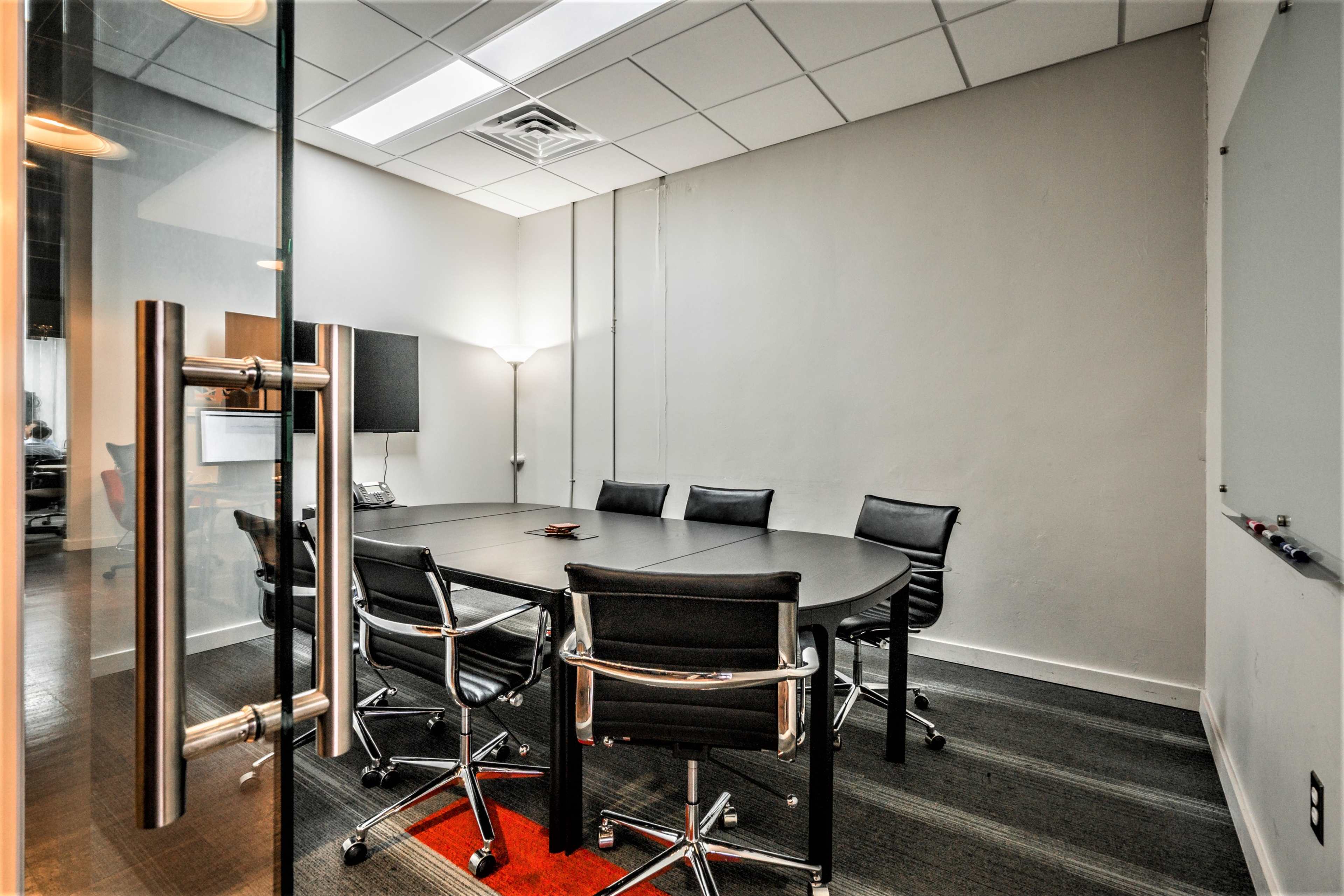 A conference room features a large round table surrounded by black chairs, with a television mounted on the wall and a lamp in the corner.