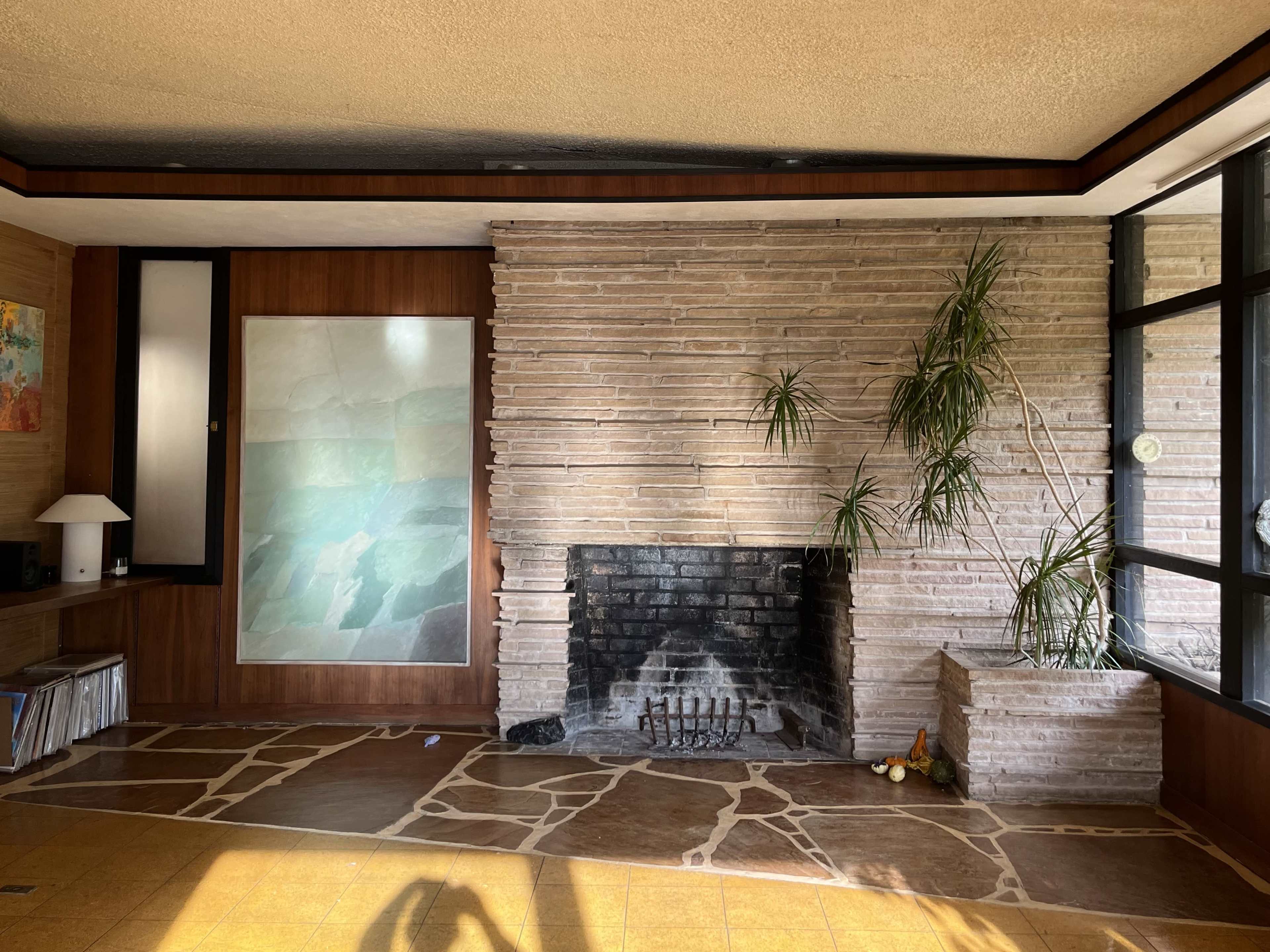 The image shows a spacious living room with a stone fireplace, a large window, and a textured wall featuring a painted panel.