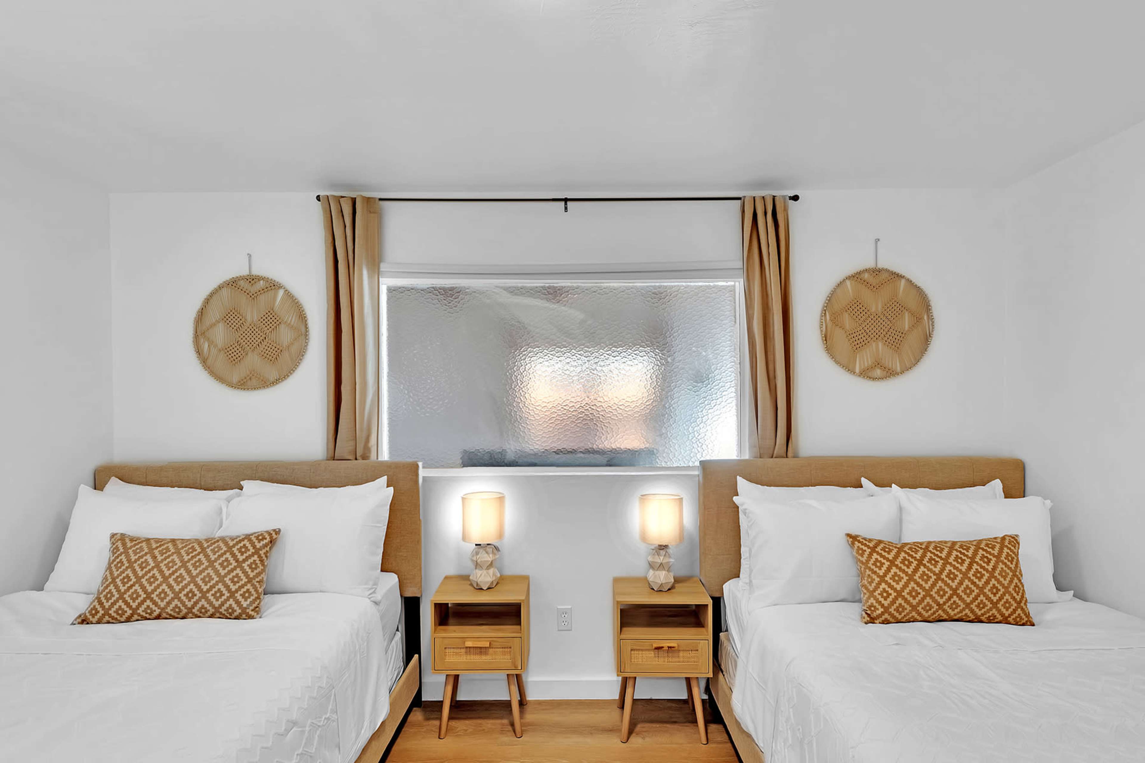 A bright bedroom with two twin beds, each paired with a bedside table and lamp, and a large window with textured glass in the background.